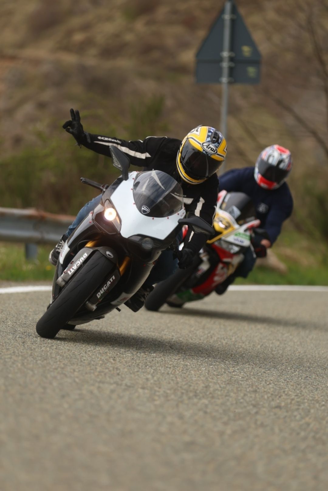 Two motorcyclists racing on a track, with the lead Ducati rider leaning into a curve while gesturing with one hand.