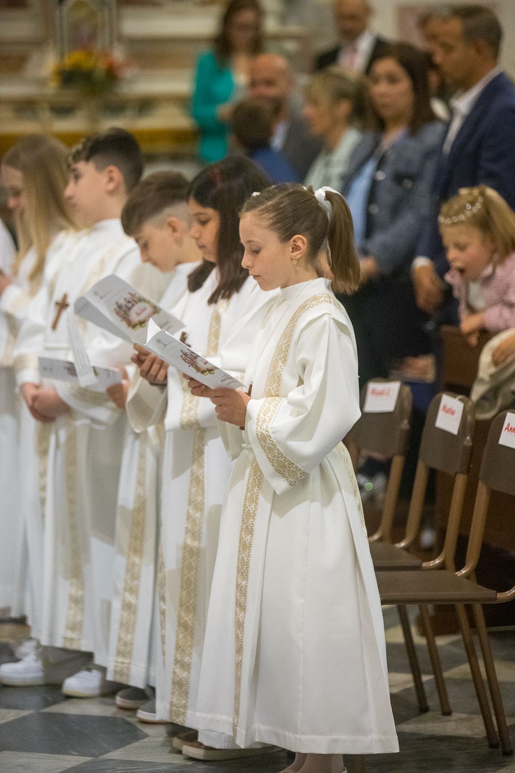 Children in white robes holding prayer books during religious service.