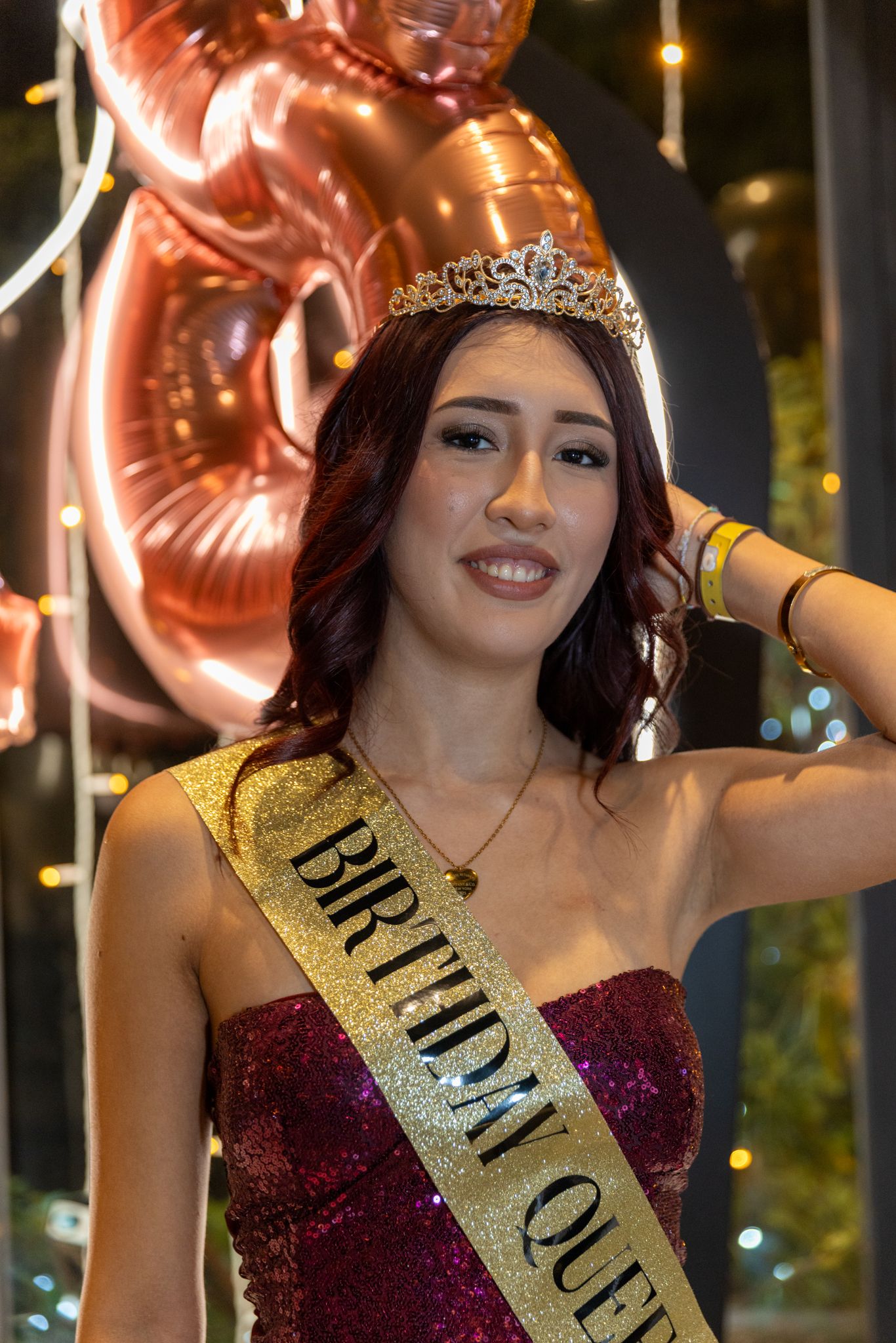 Beauty pageant contestant in burgundy gown and gold sash poses with celebratory decorations and lights in background.