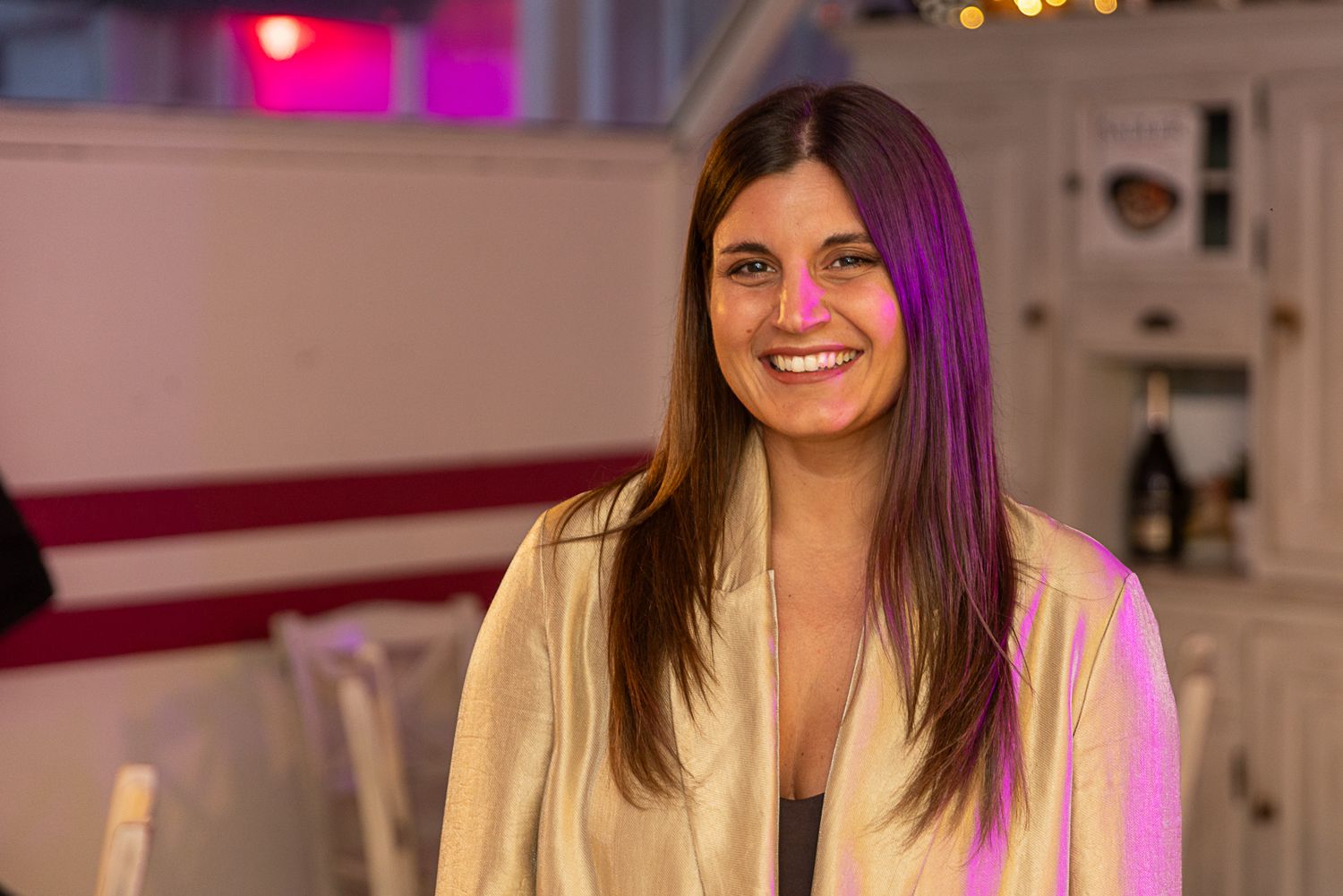 Smiling woman with long brown hair in a golden blazer under colorful purple and pink lighting at an indoor event.