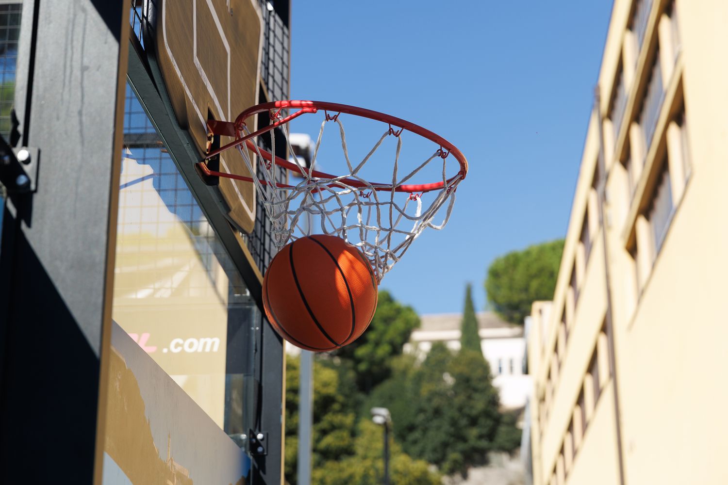 Basketball players practicing shots at an outdoor court with multiple hoops mounted on a building wall.