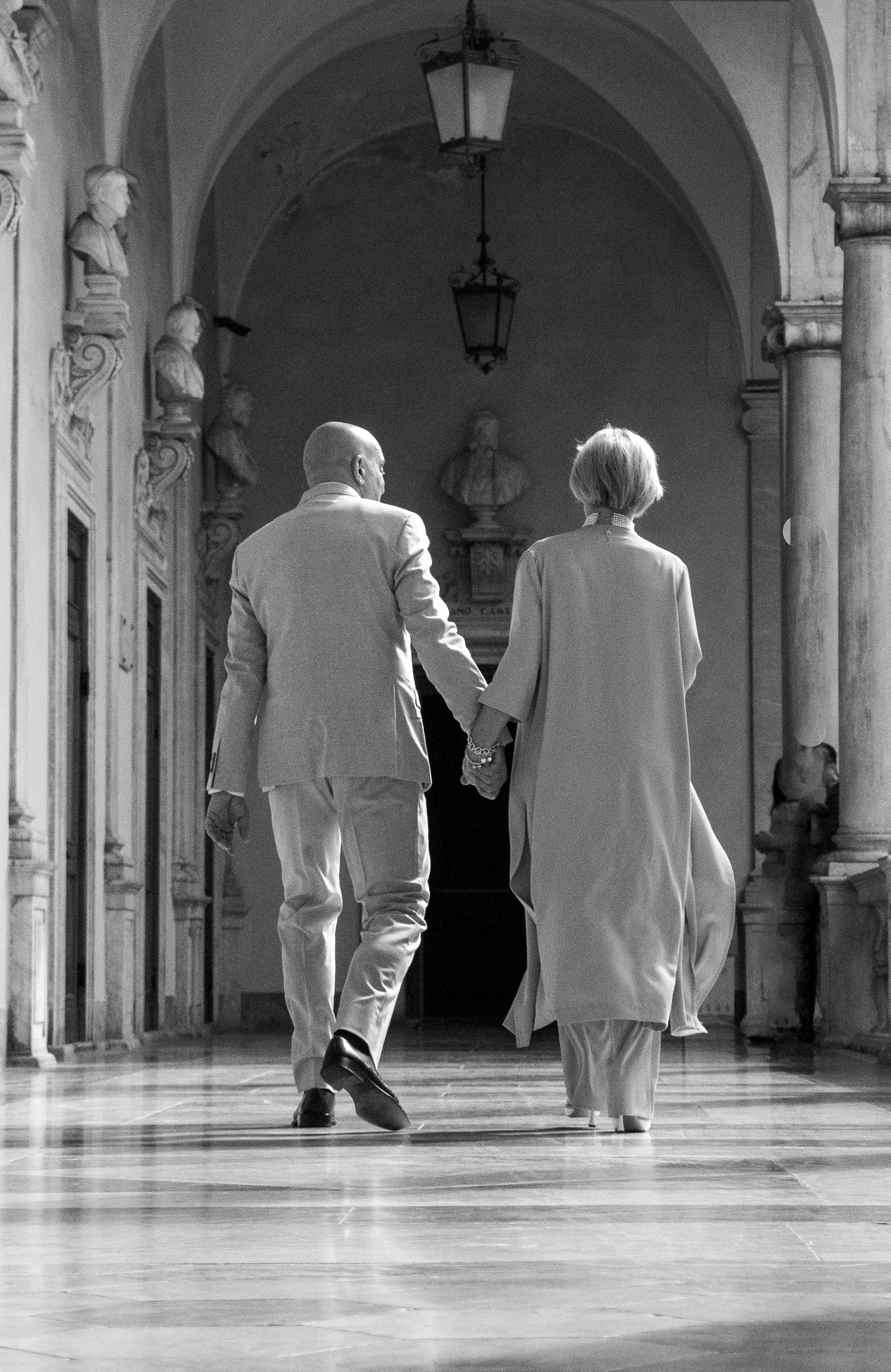 Black and white image of people walking through an arched corridor with classical architecture.