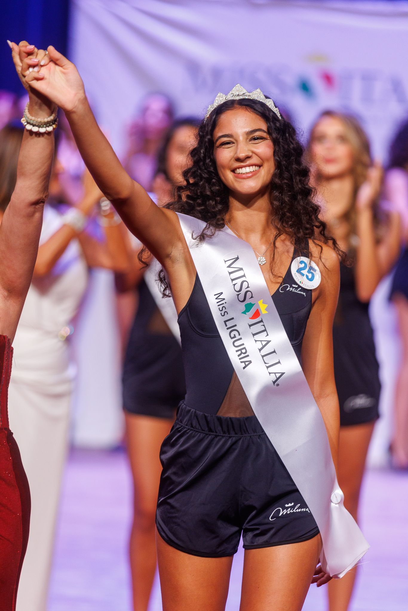 Pageant winner celebrates with raised arm wearing sash and crown at Miss Italia competition.