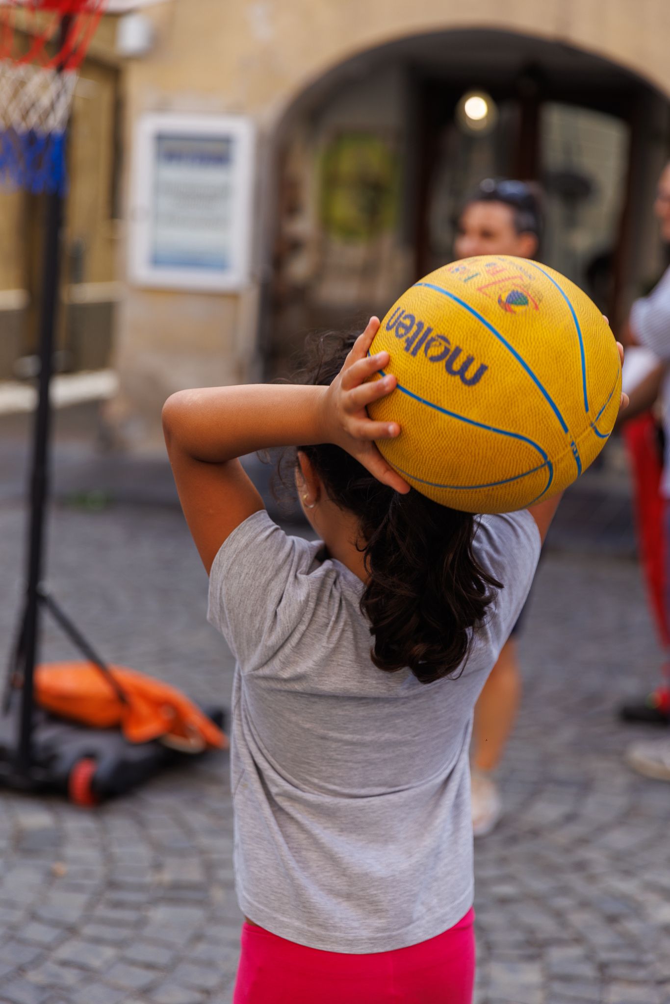 Child playing with yellow basketball while wearing gray t-shirt on cobblestone street.