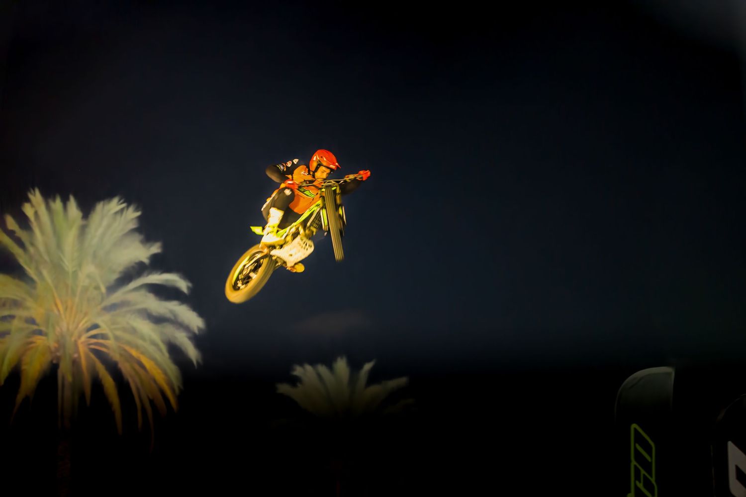 Silhouette of a motorcycle performing a jump against a dark sky with palm trees.