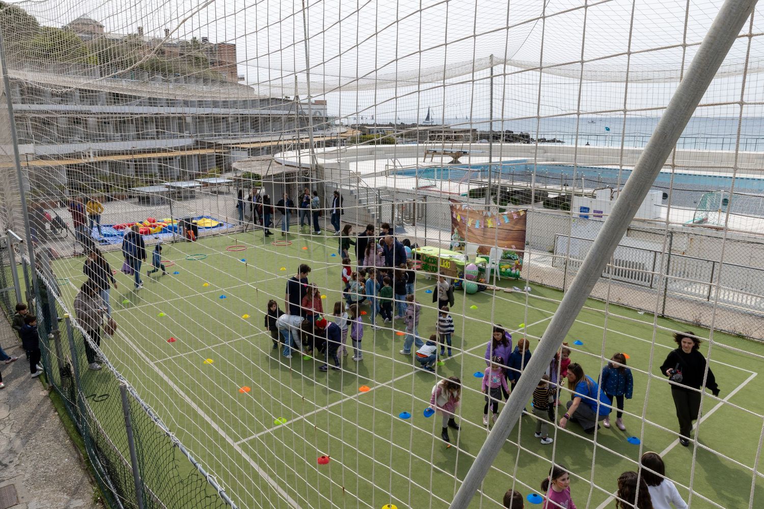 Children playing on an outdoor artificial turf soccer field near a coastal building with ocean views.