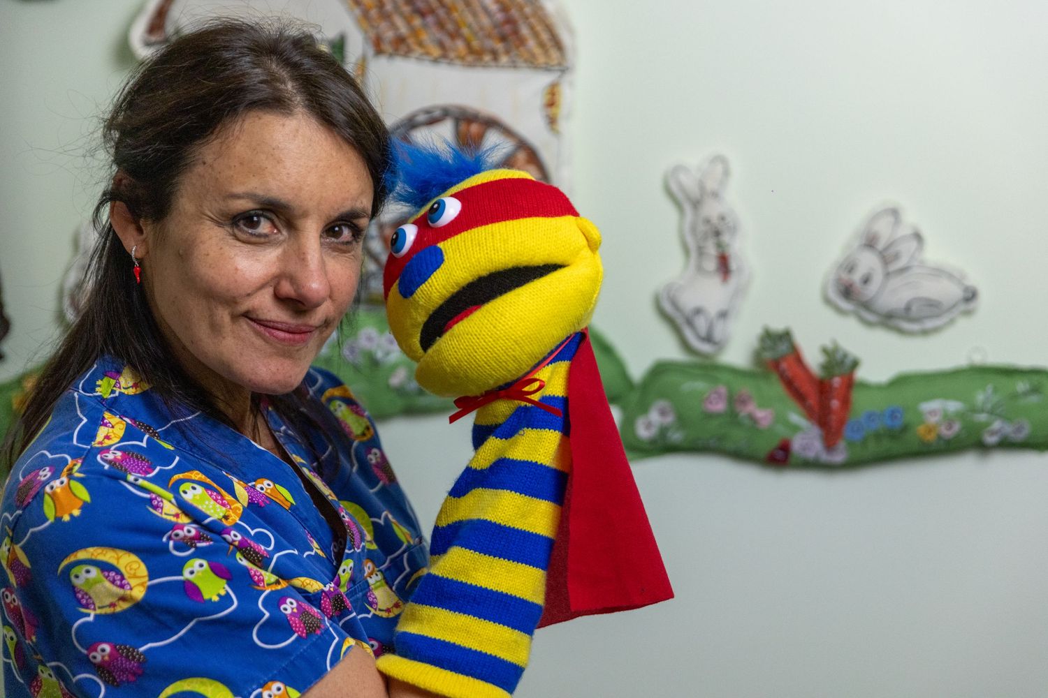 A colorful sock puppet with yellow and blue stripes is shown against a wall with children's drawings.