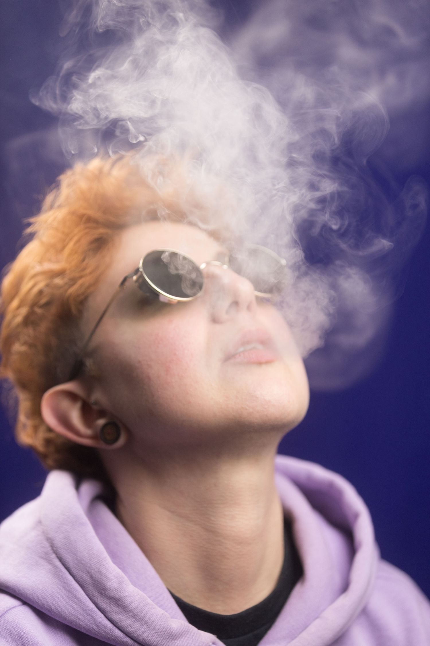 Person in purple hoodie exhaling smoke against blue background while wearing sunglasses.