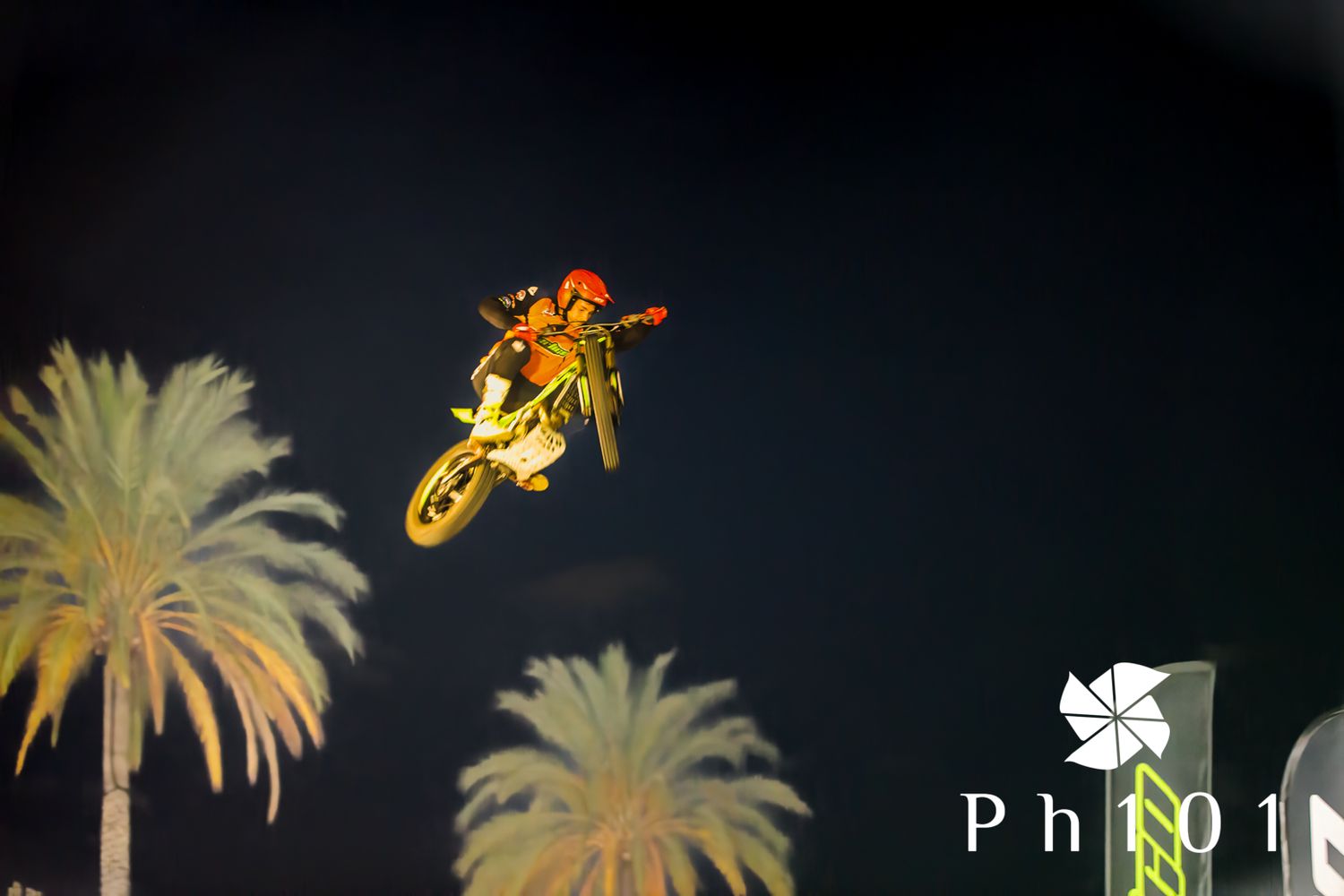 Motorcycle performing a dramatic jump against a night sky with palm trees.