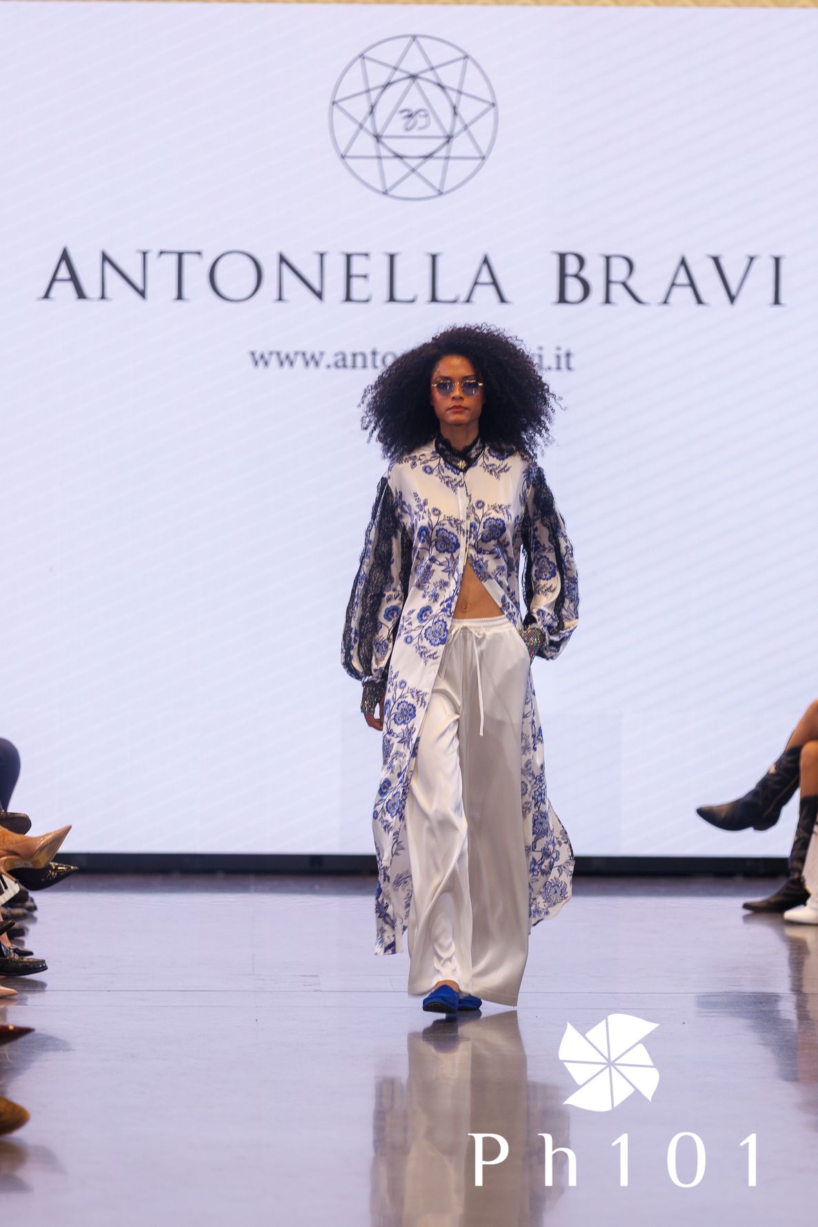 Fashion model presents elegant white pants and printed jacket on runway for Antonella Bravi collection.