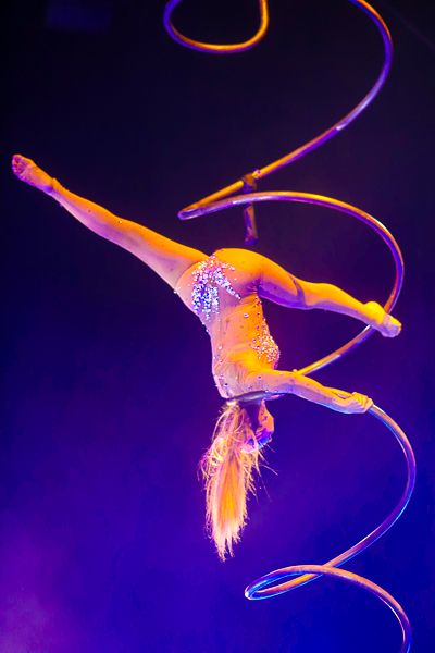 Aerial acrobat performing with ribbons against purple and blue lighting.