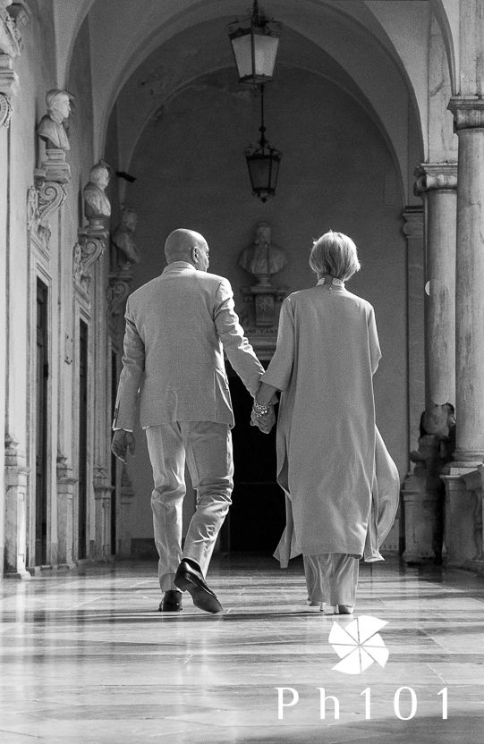 Black and white photo of a couple walking hand in hand through an elegant arched corridor.