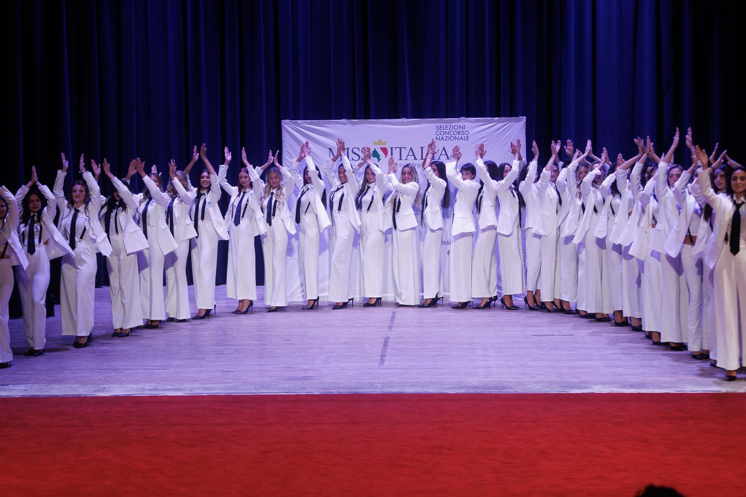 Large choir group in white robes performs on stage with red carpet and blue backdrop.