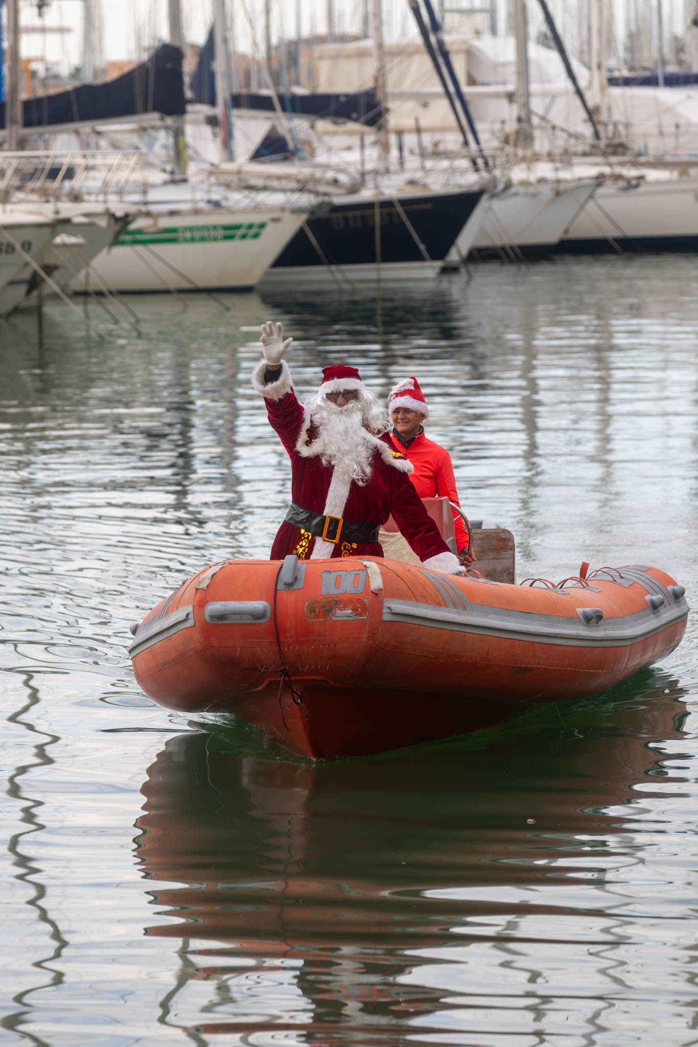 Santa Claus riding in an orange inflatable rescue boat through a marina.