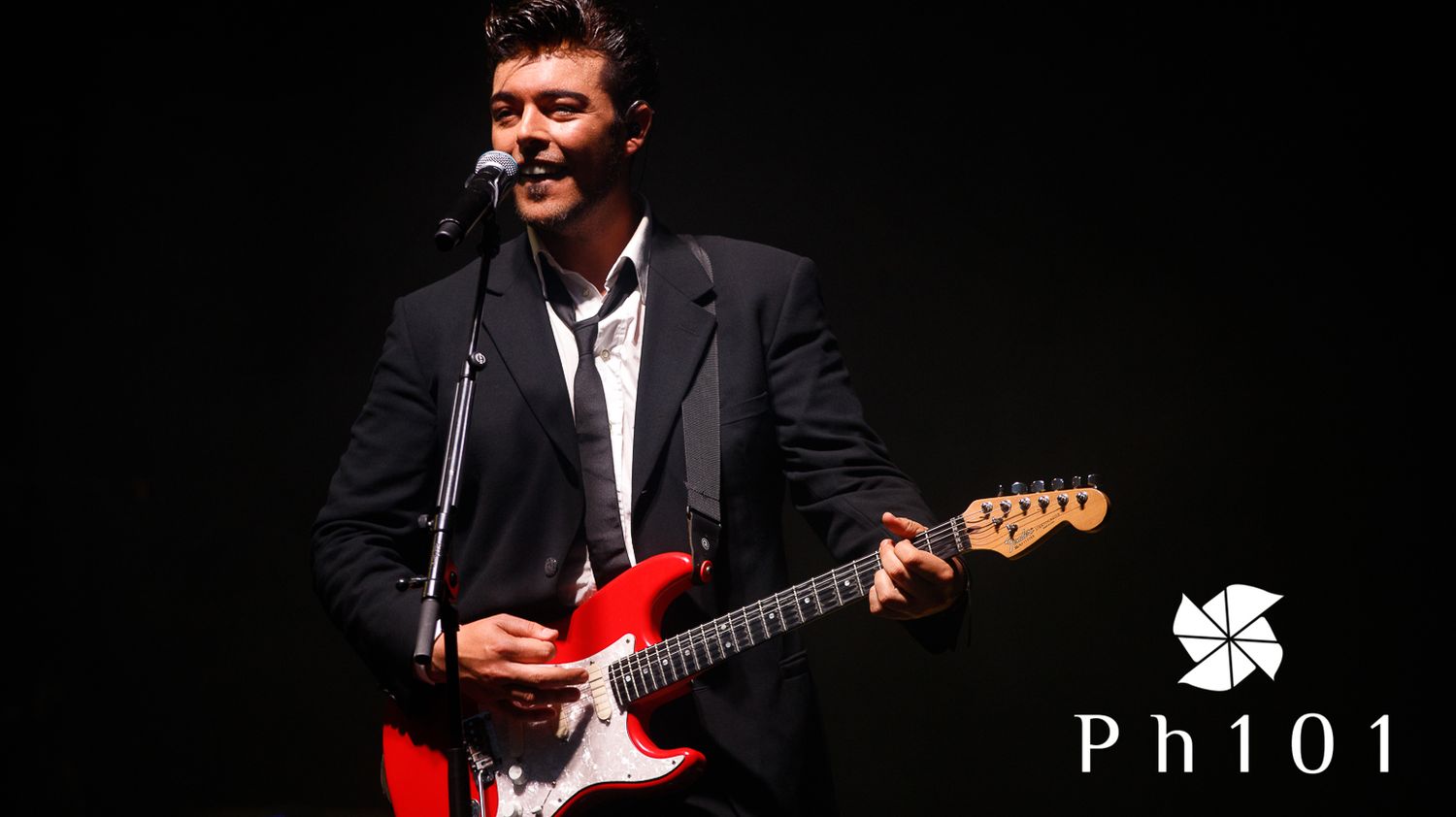 Musicians performing on stage with guitars against black background.