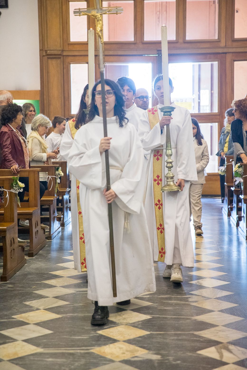 People in white robes carrying crosses process through church with checkered floor.