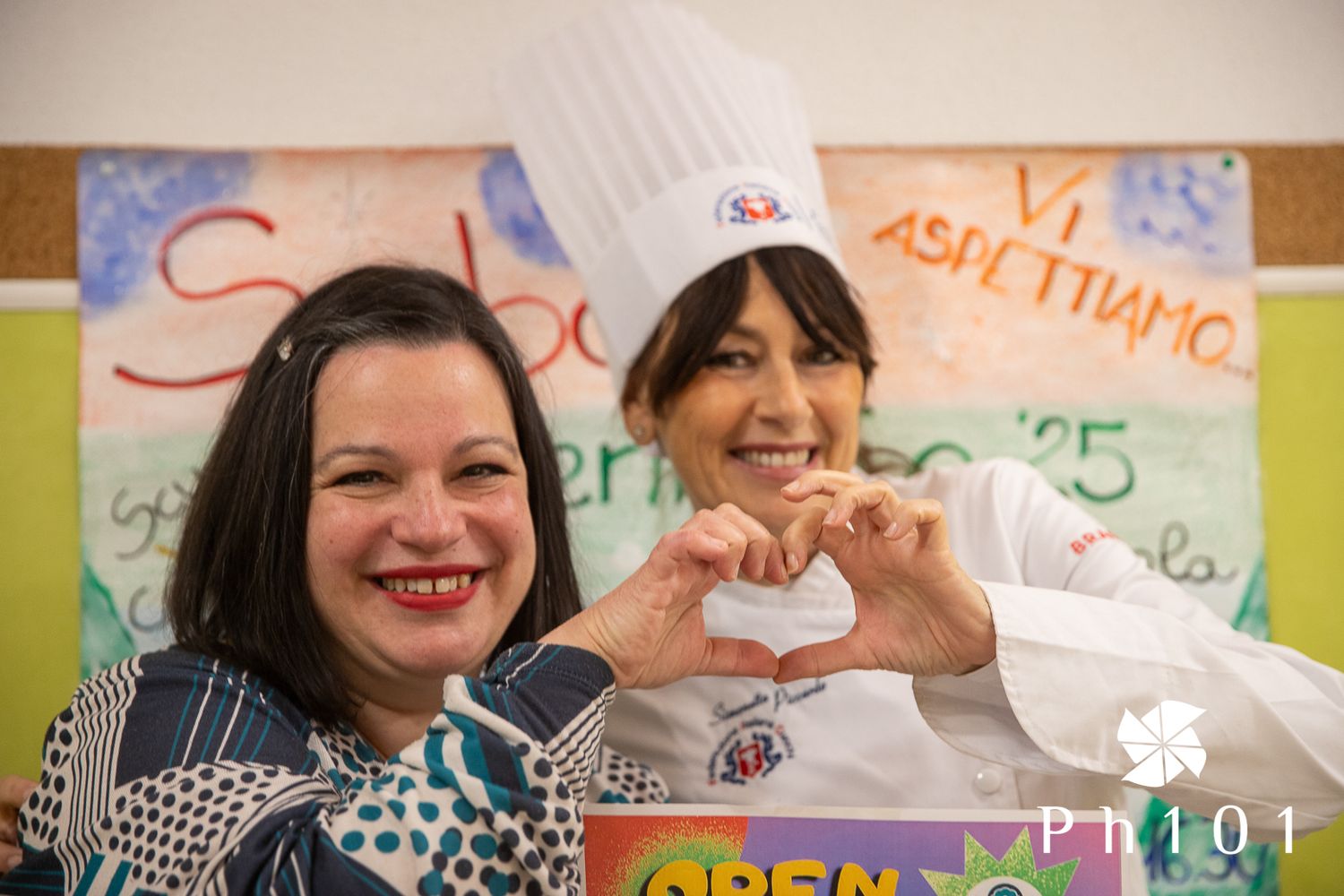 Two educators making heart shapes with their hands in front of a colorful bulletin board.