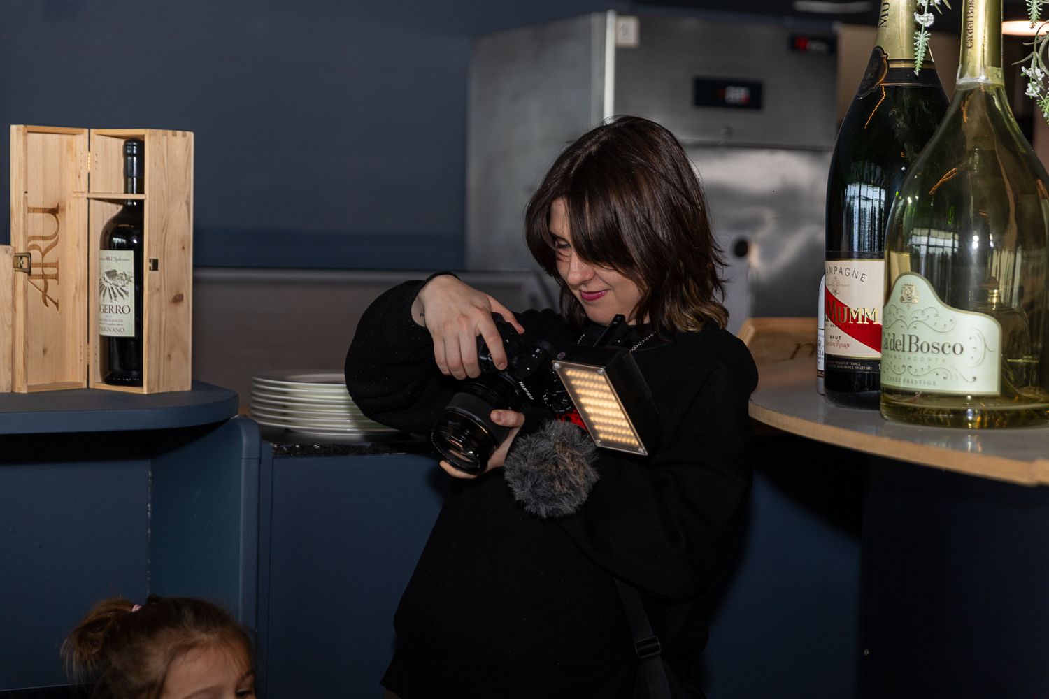 Woman in black outfit holding a camera near wine bottles and a wooden box in a dimly lit venue.