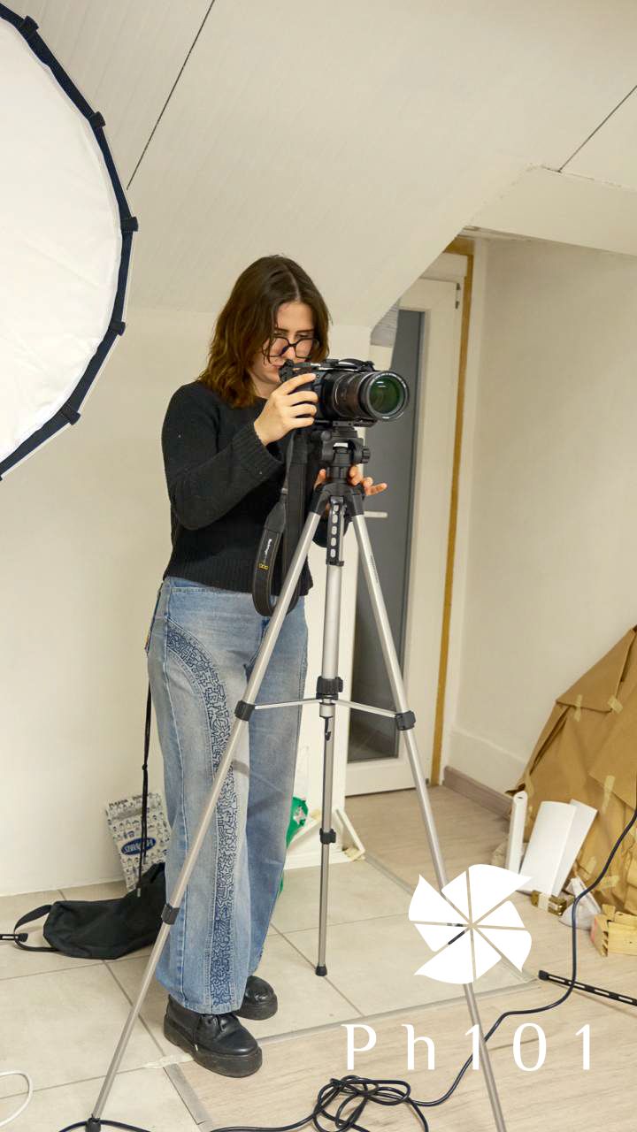 Photographer in black sweater and jeans crouches with camera on tripod in studio with lighting equipment.