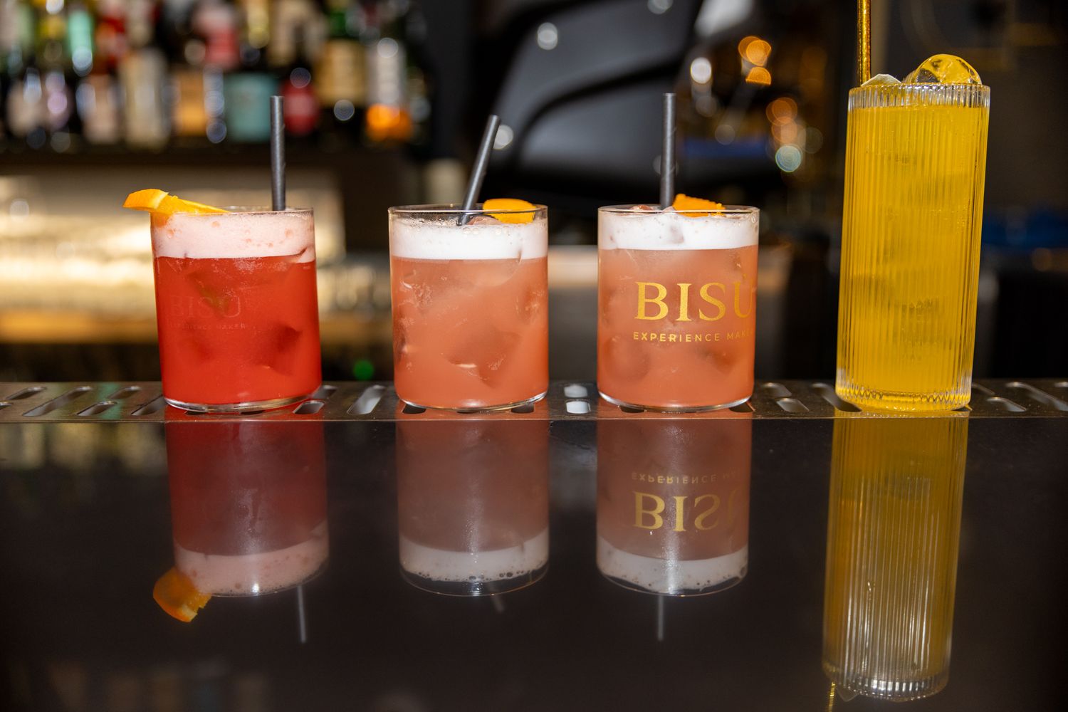A row of colorful craft cocktails in glasses lined up on a dark bar counter with ambient lighting.