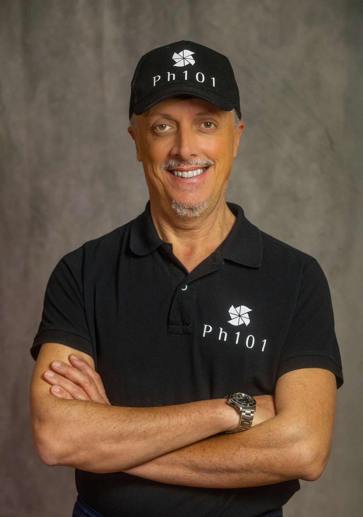 Professional headshot of someone wearing a black polo shirt and baseball cap with crossed arms against gray backdrop.