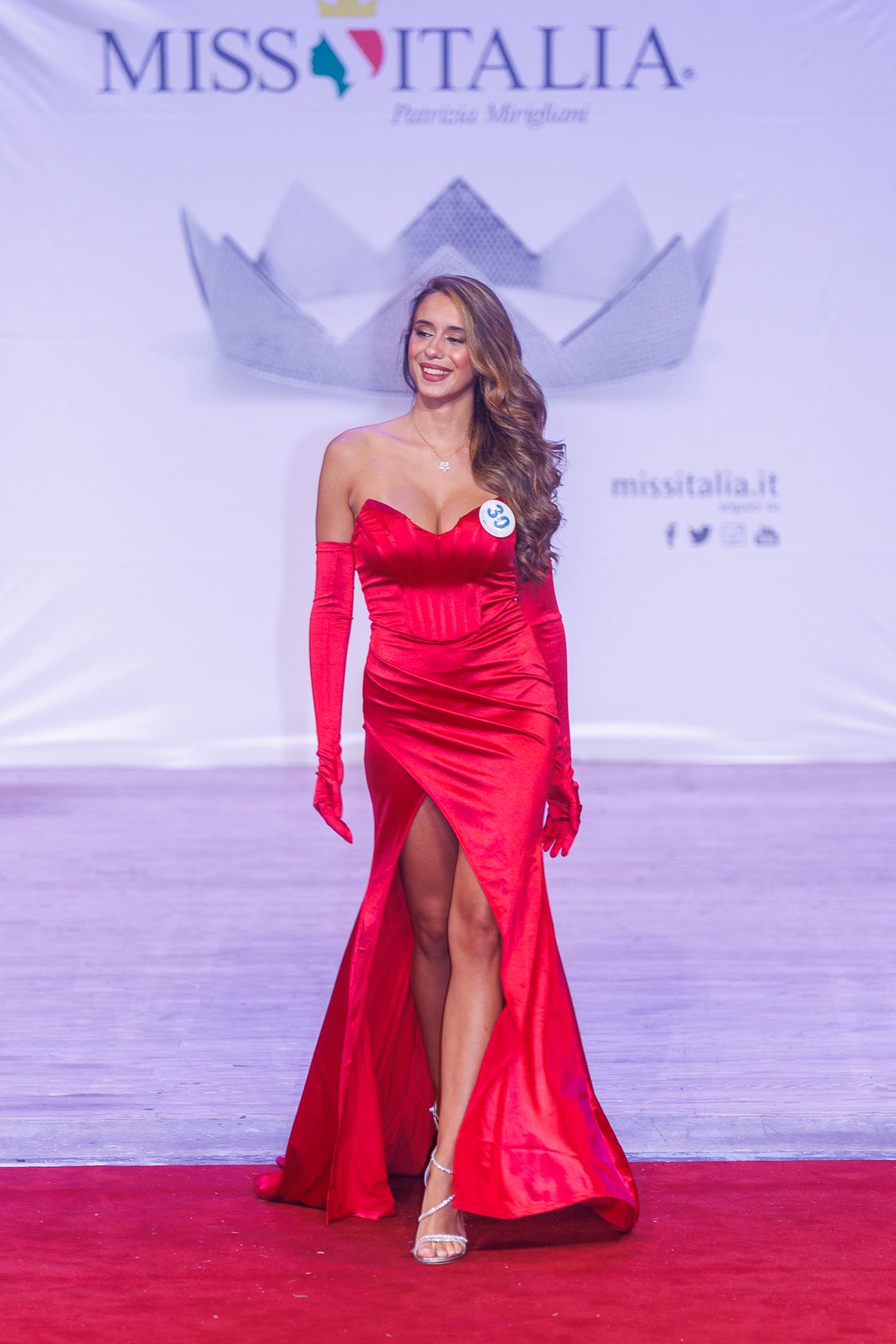 Contestant in red satin evening gown poses on Miss Italia pageant runway.