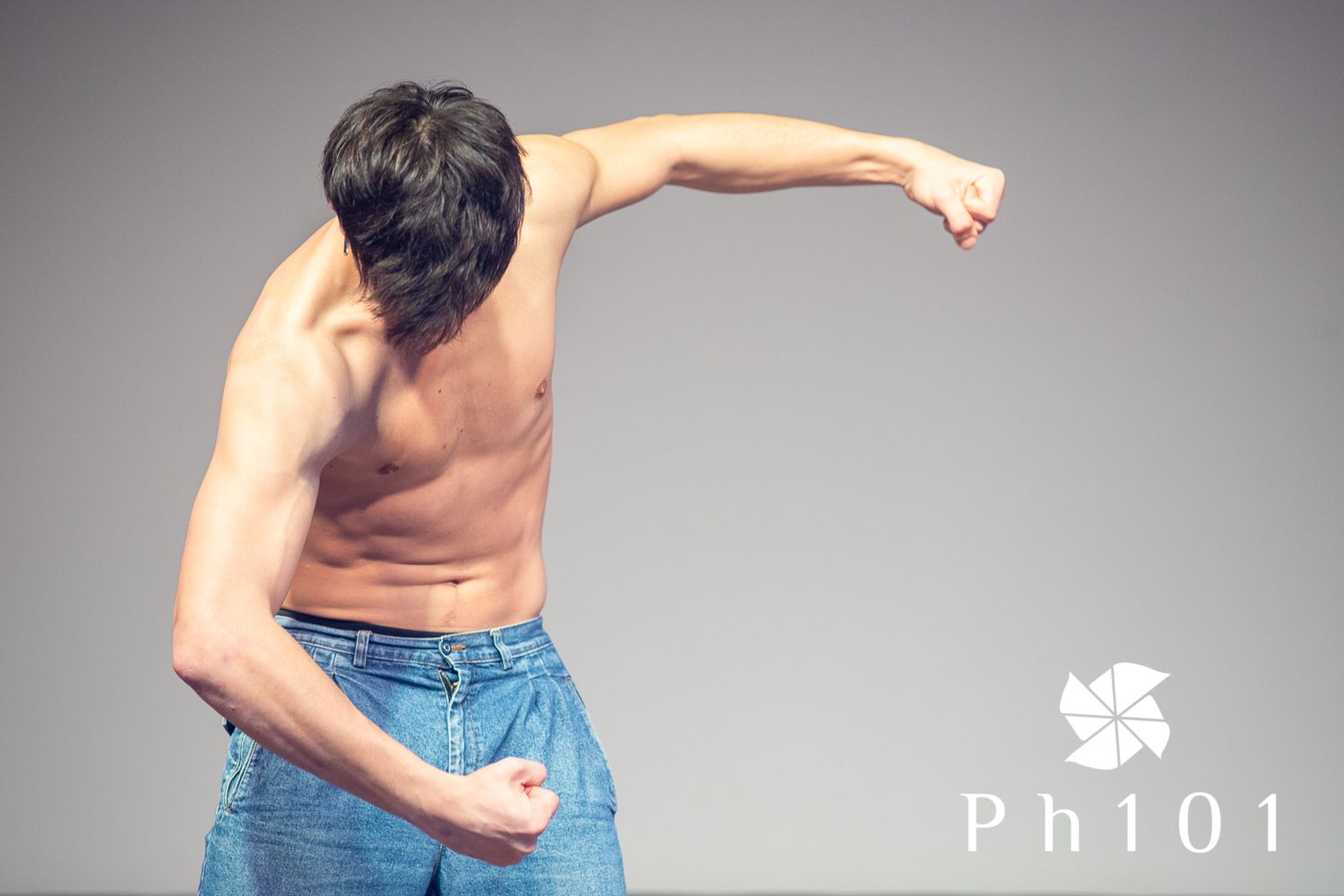 Shirtless person in jeans performing an expressive dance move against grey background.