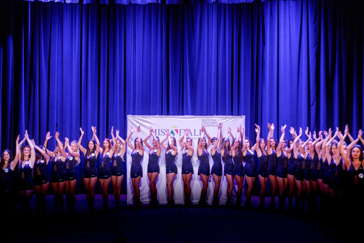 Dancers perform on stage in a synchronized line against a blue curtain backdrop.