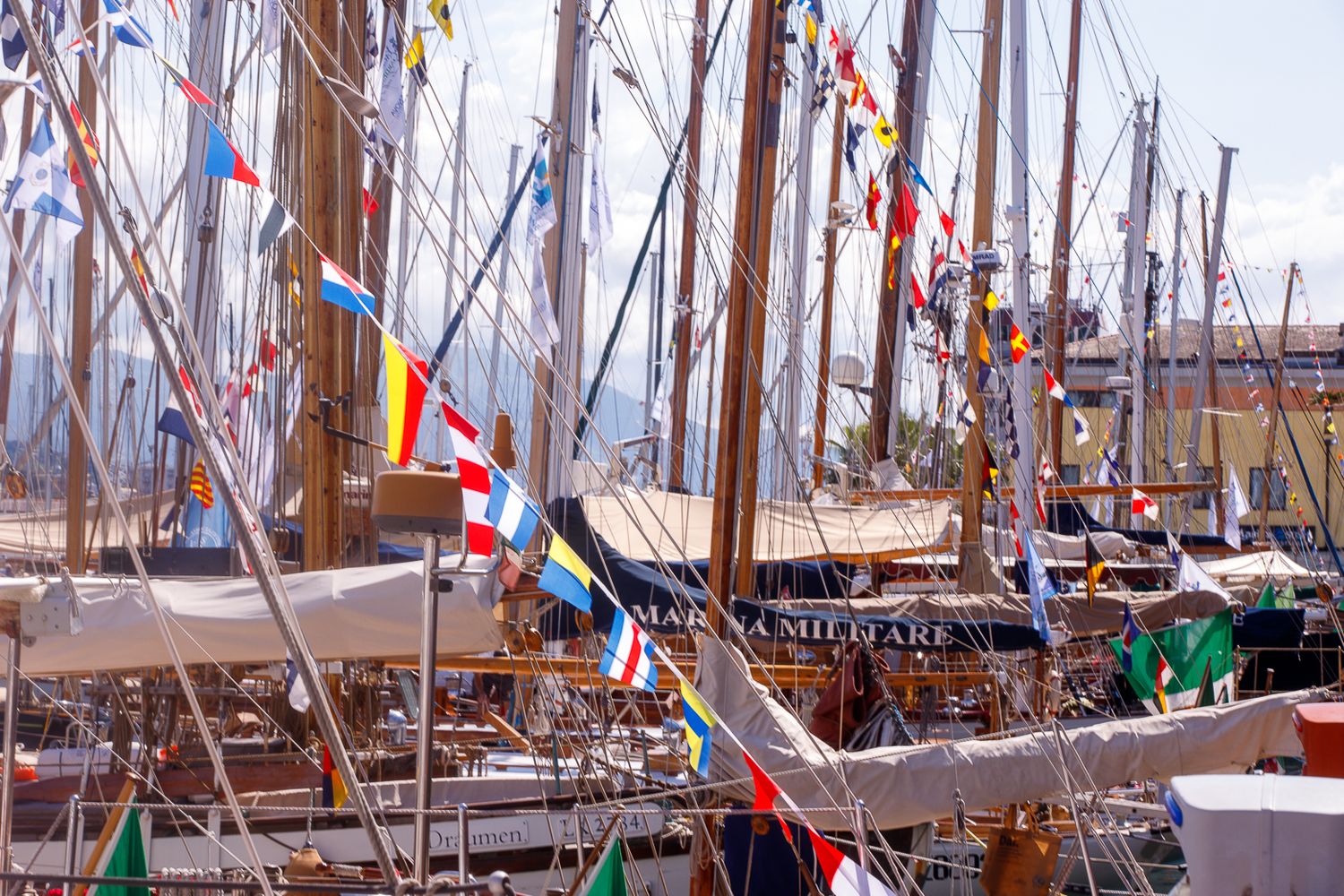 Row of sailboat masts with colorful flags and rigging against blue sky.