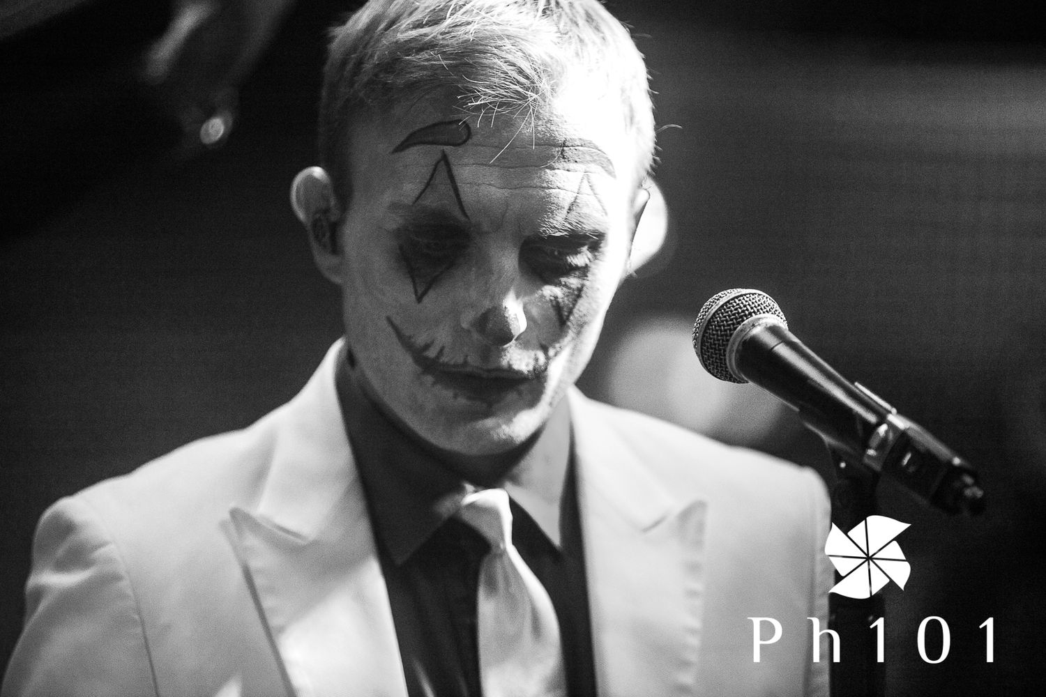 Black and white photo of a performer with face paint singing into a microphone on stage.