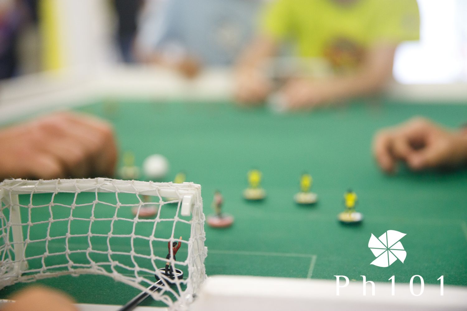 Close-up view of a miniature soccer goal on a green gaming table with players in the background.