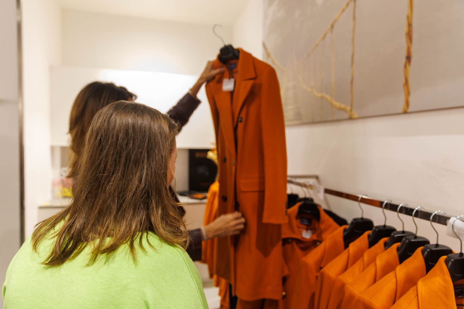 Person in neon yellow shirt examining orange coat on retail clothing rack.