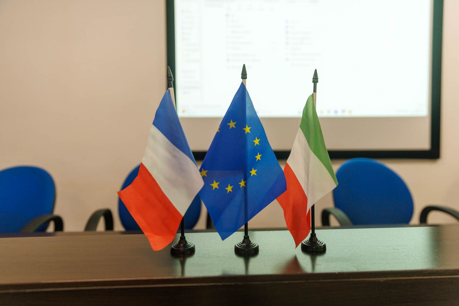 French, European Union, and Italian flags displayed on desk in meeting room.