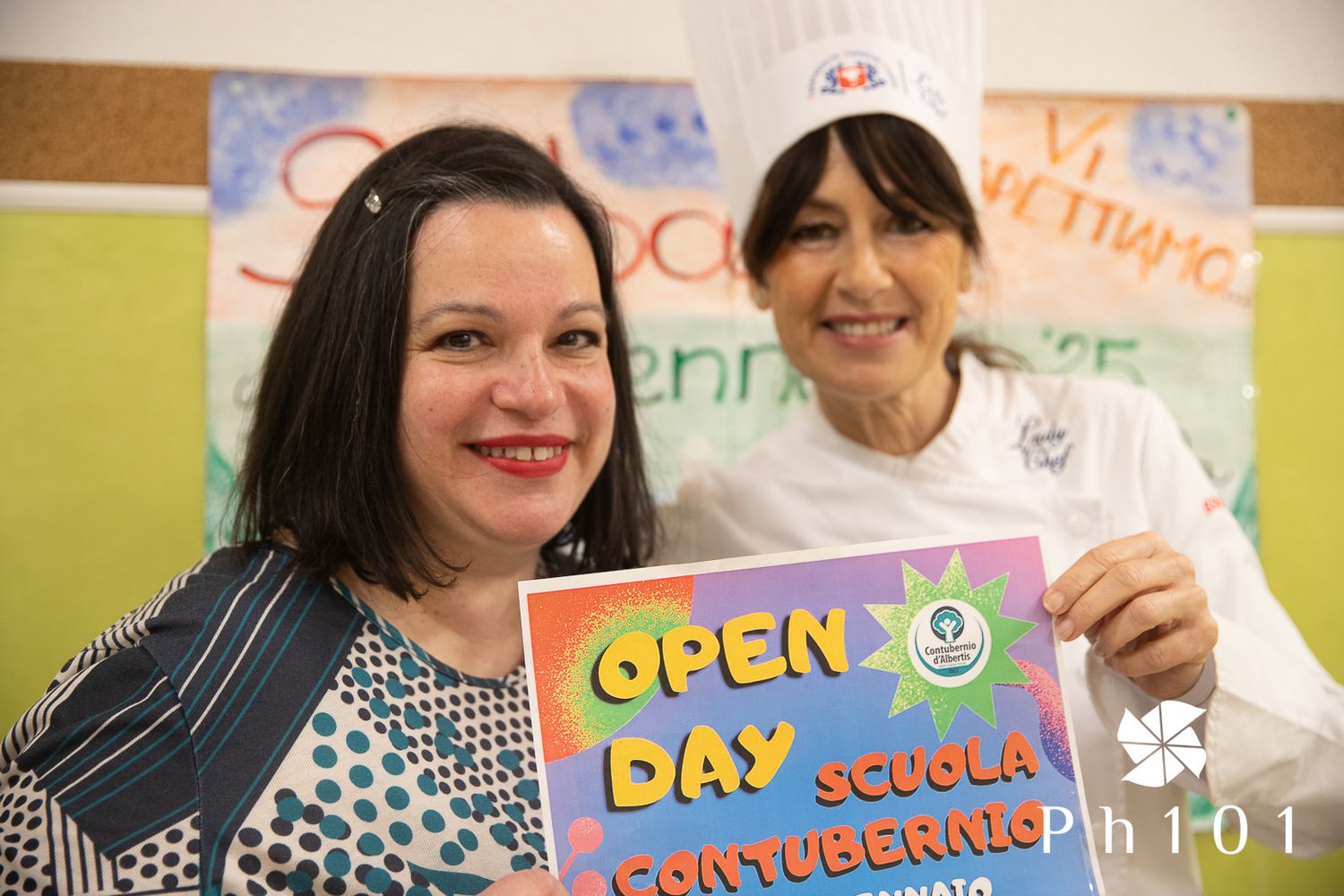 Two educators holding an Open Day sign in a classroom with colorful posters on the wall.