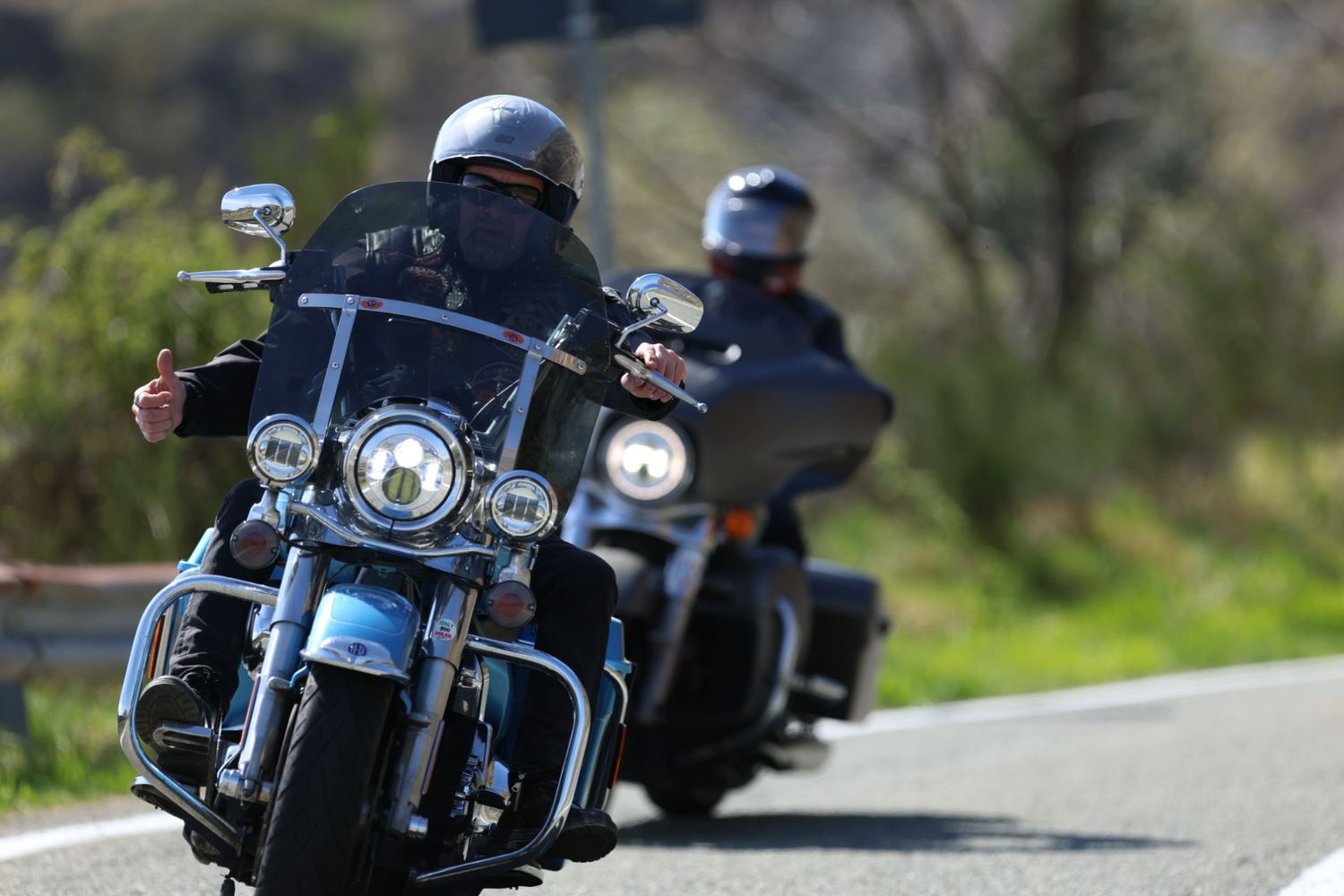 Two motorcyclists riding Harley-Davidson touring bikes on a scenic road on a sunny day.