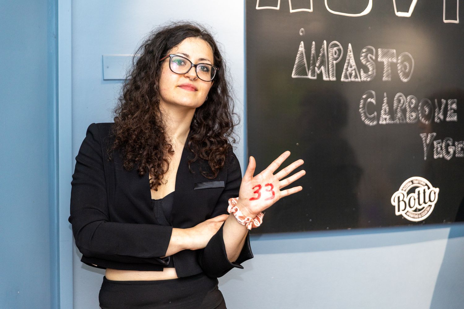 A person in black attire gestures while standing next to a chalkboard with writing in a casual business setting.