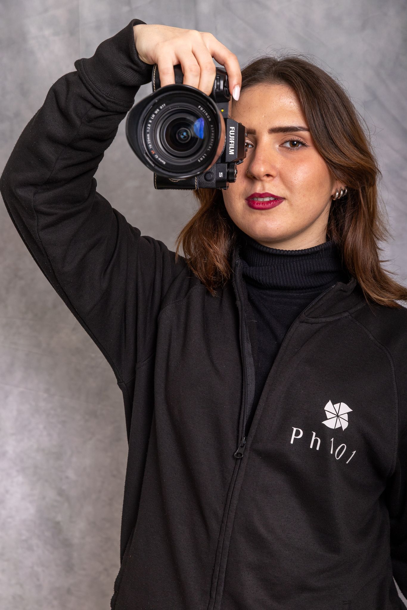 A photographer wearing a black hoodie holds up a DSLR camera in various poses against a gray studio background.