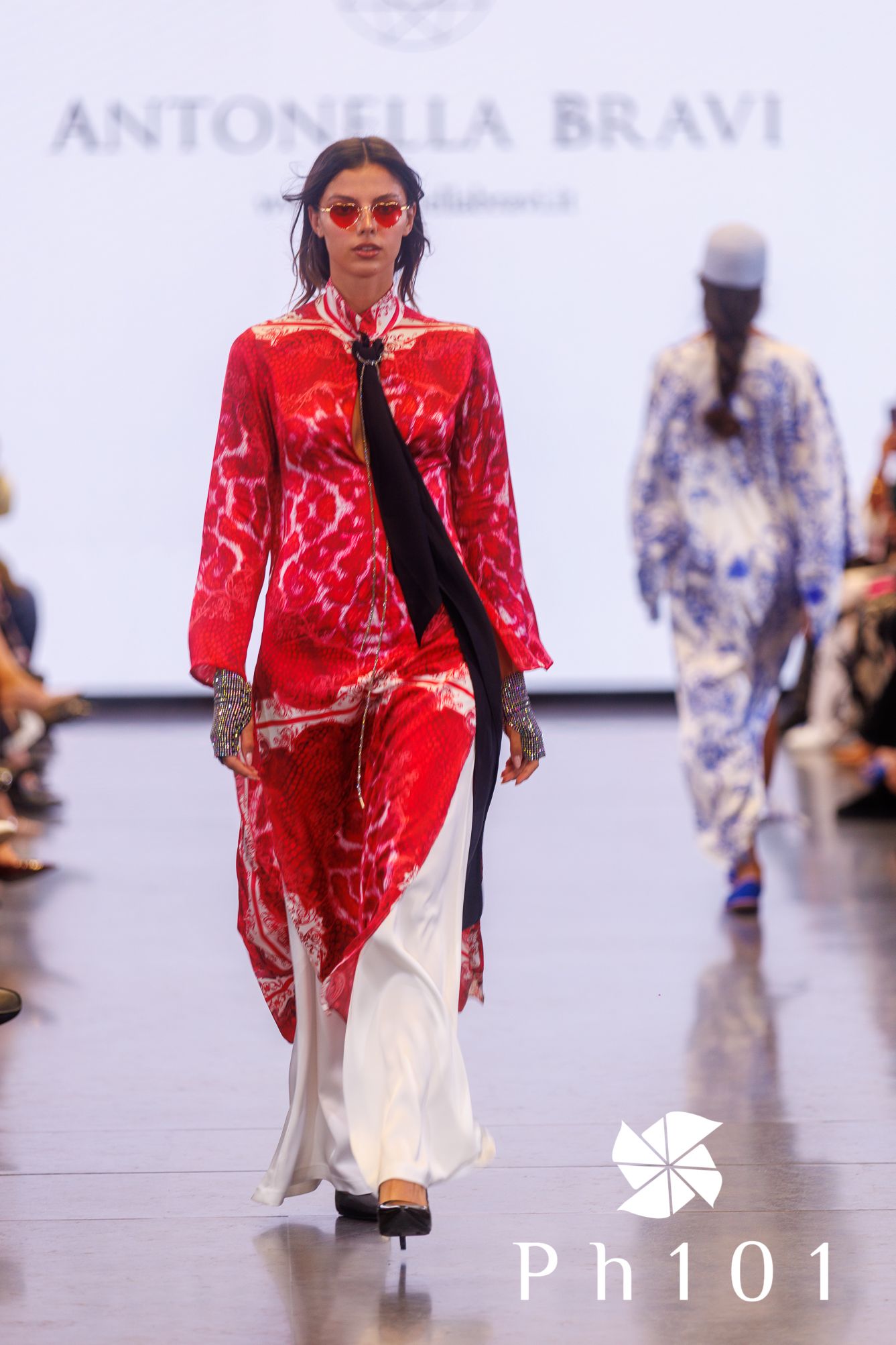 Fashion model walks runway in vibrant red patterned jacket and white dress at Antonia Bravi fashion show.