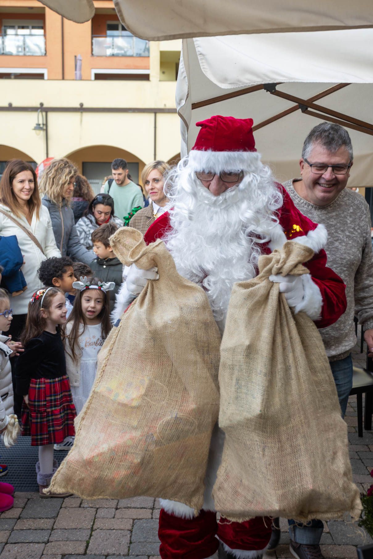 Santa Claus displaying large burlap gift sacks to excited children at a holiday event.