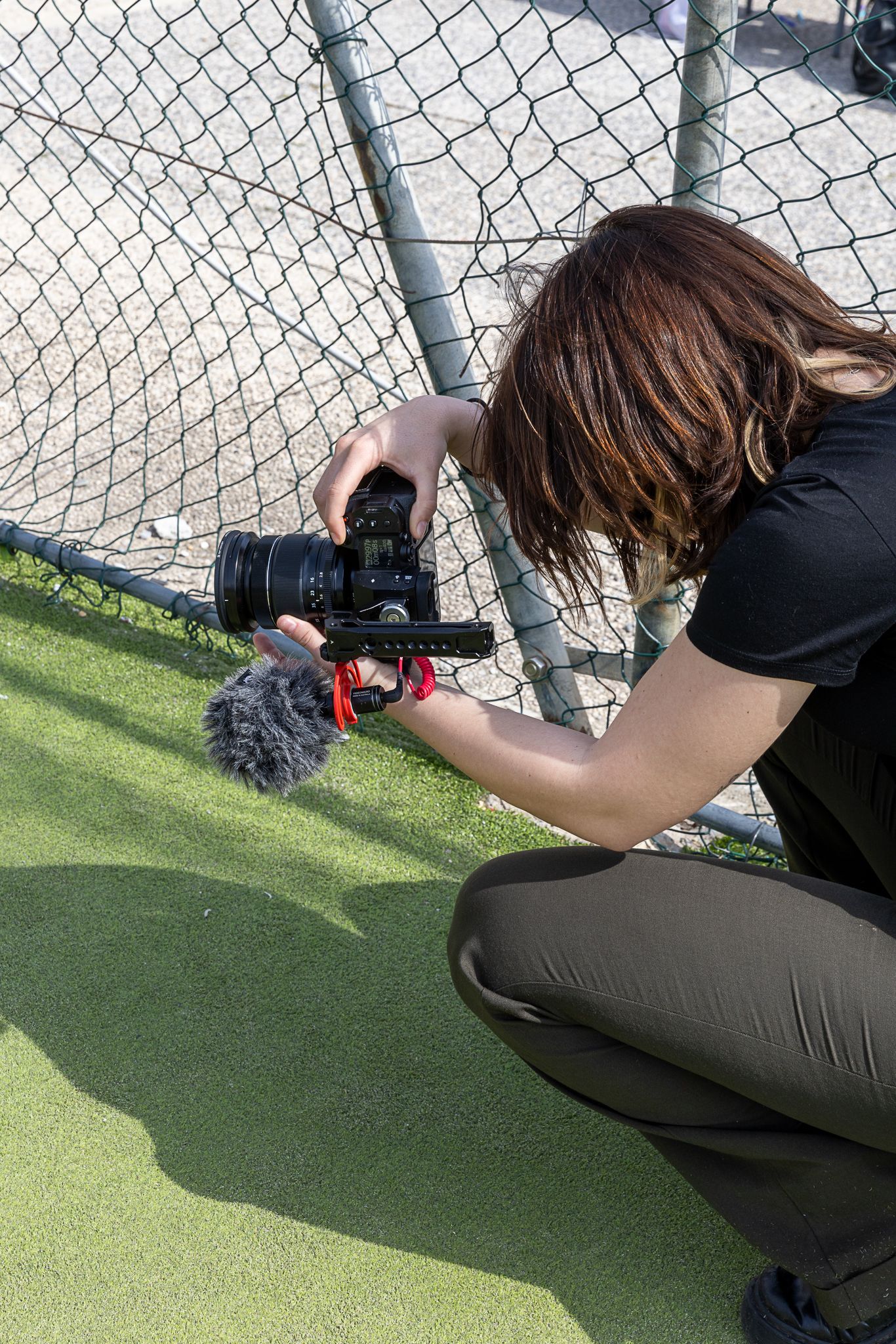 Photographer crouching on artificial turf, filming with a DSLR camera and microphone near a chain-link fence outdoors.