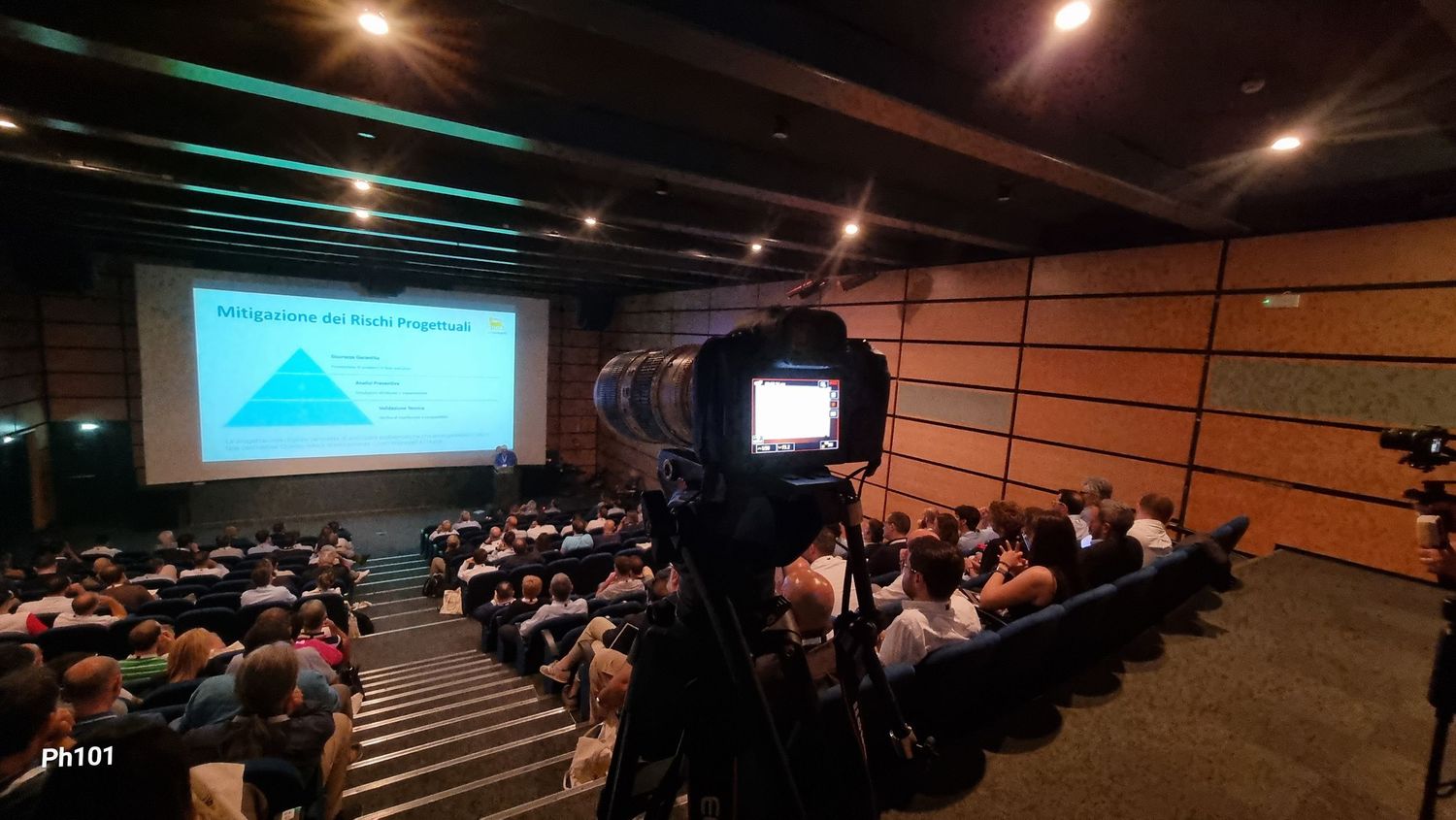 Wide angle view of filled auditorium with blue lighting and presentation screen.