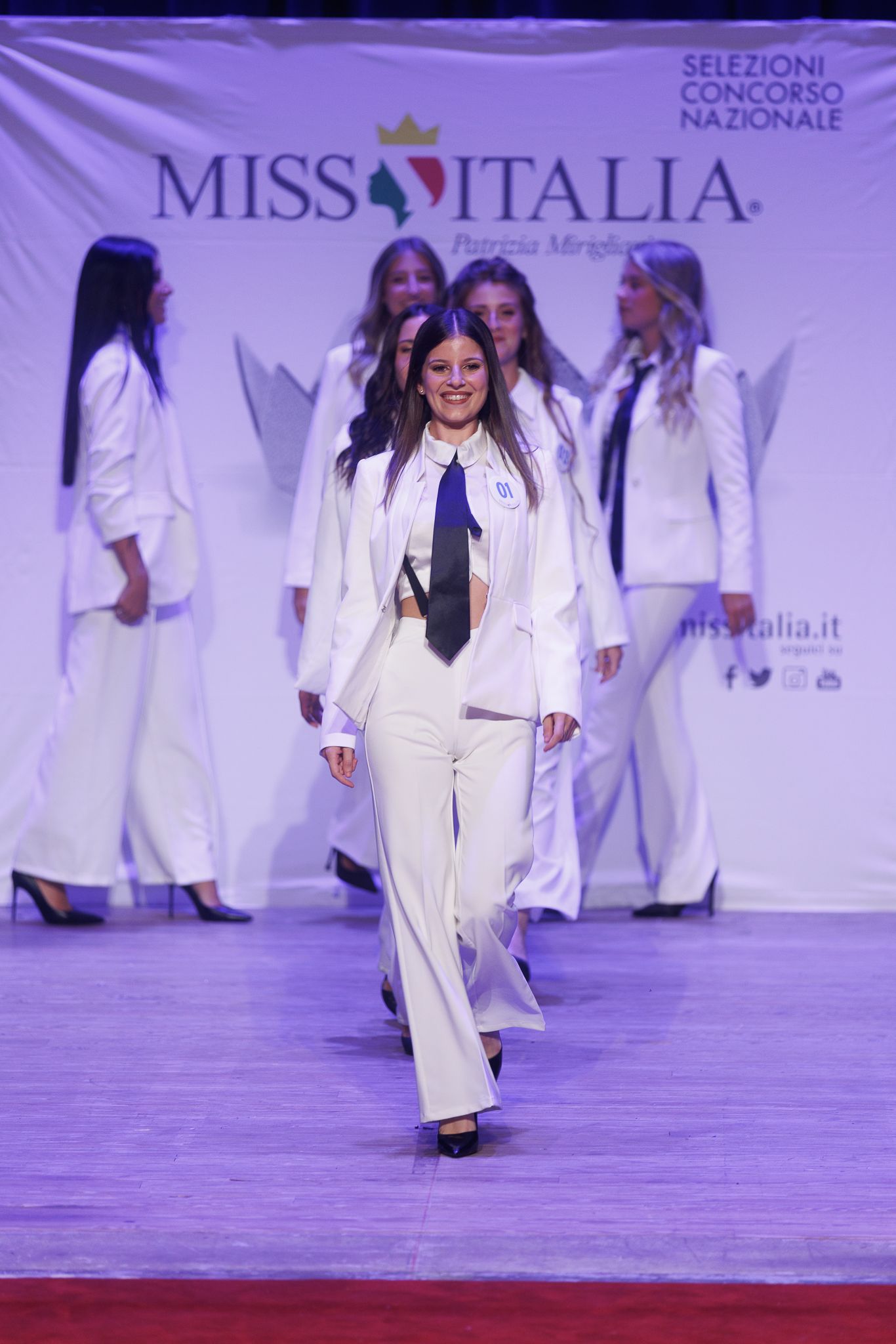 Model walks runway in white pantsuit during Miss Italia fashion show.