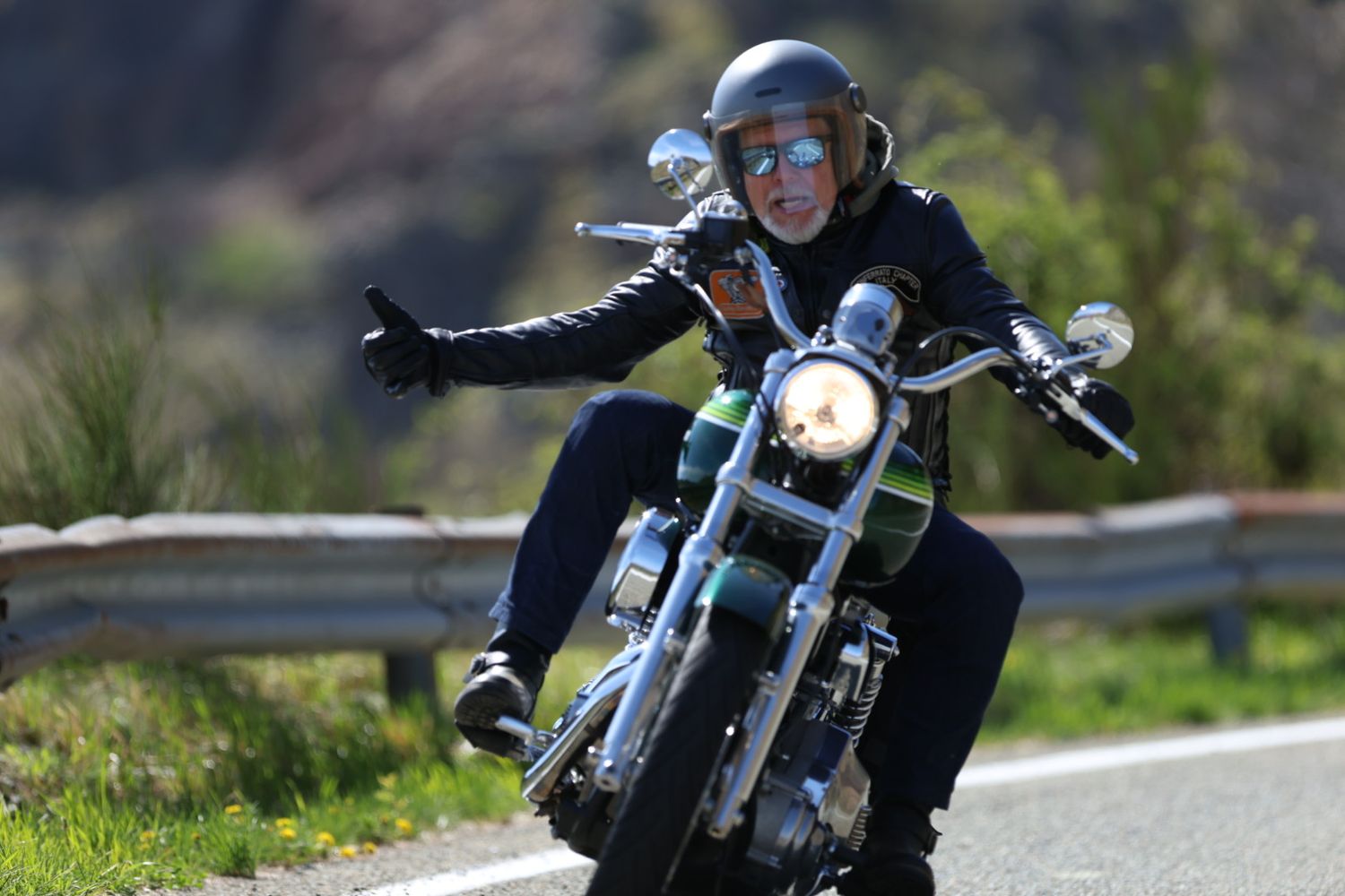 A motorcyclist in a helmet and sunglasses rides a classic cruiser motorcycle on a sunny road.