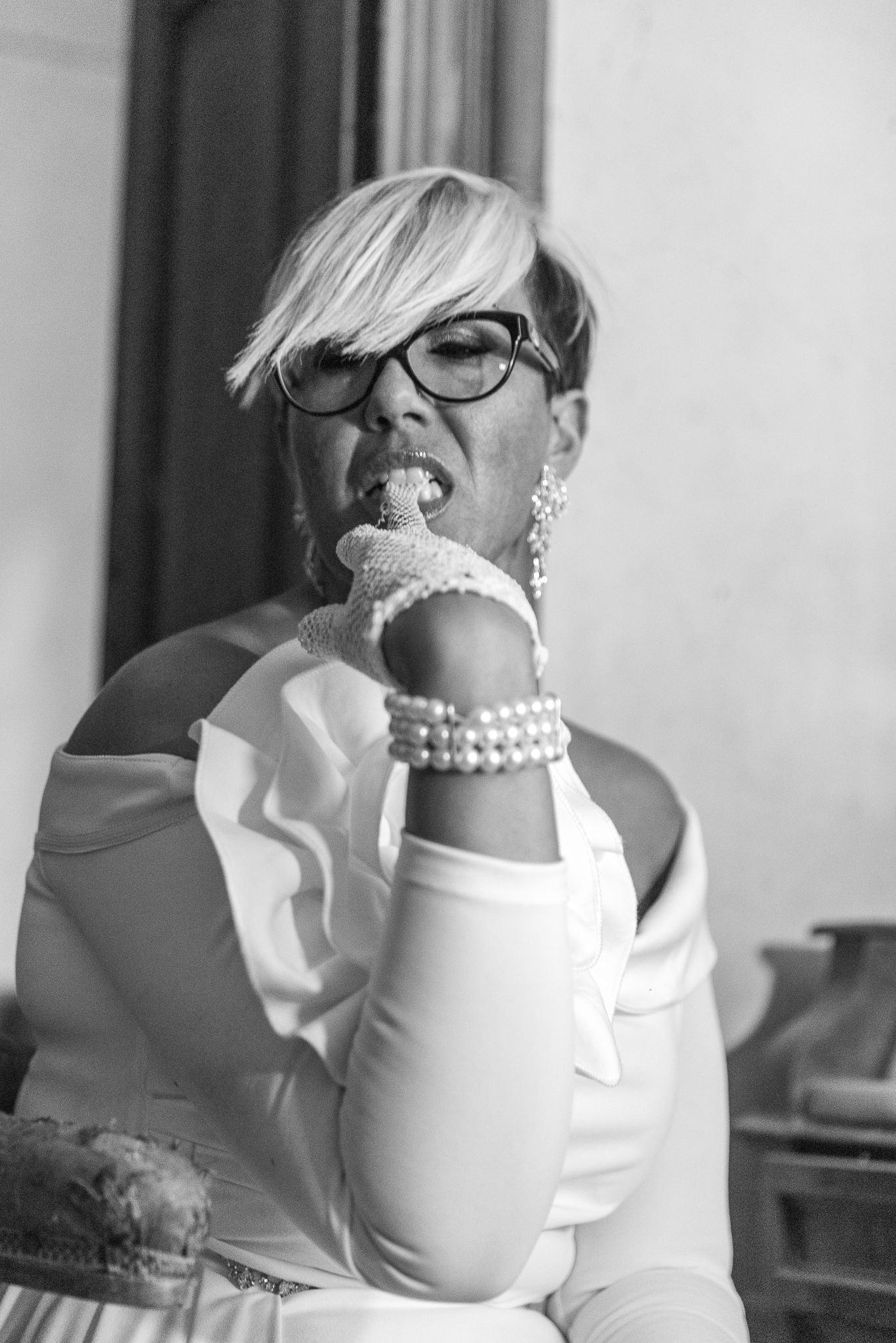 Bride in white dress with pearl accessories and glasses poses playfully in black and white portrait.
