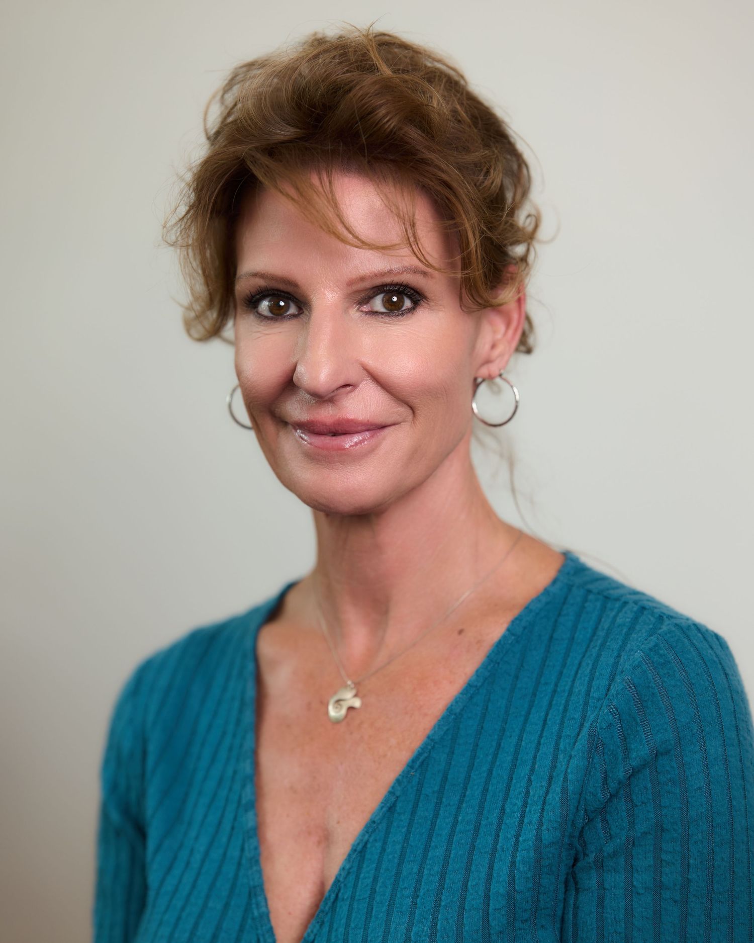 Professional headshot of a smiling woman with auburn hair wearing a teal sweater and hoop earrings.