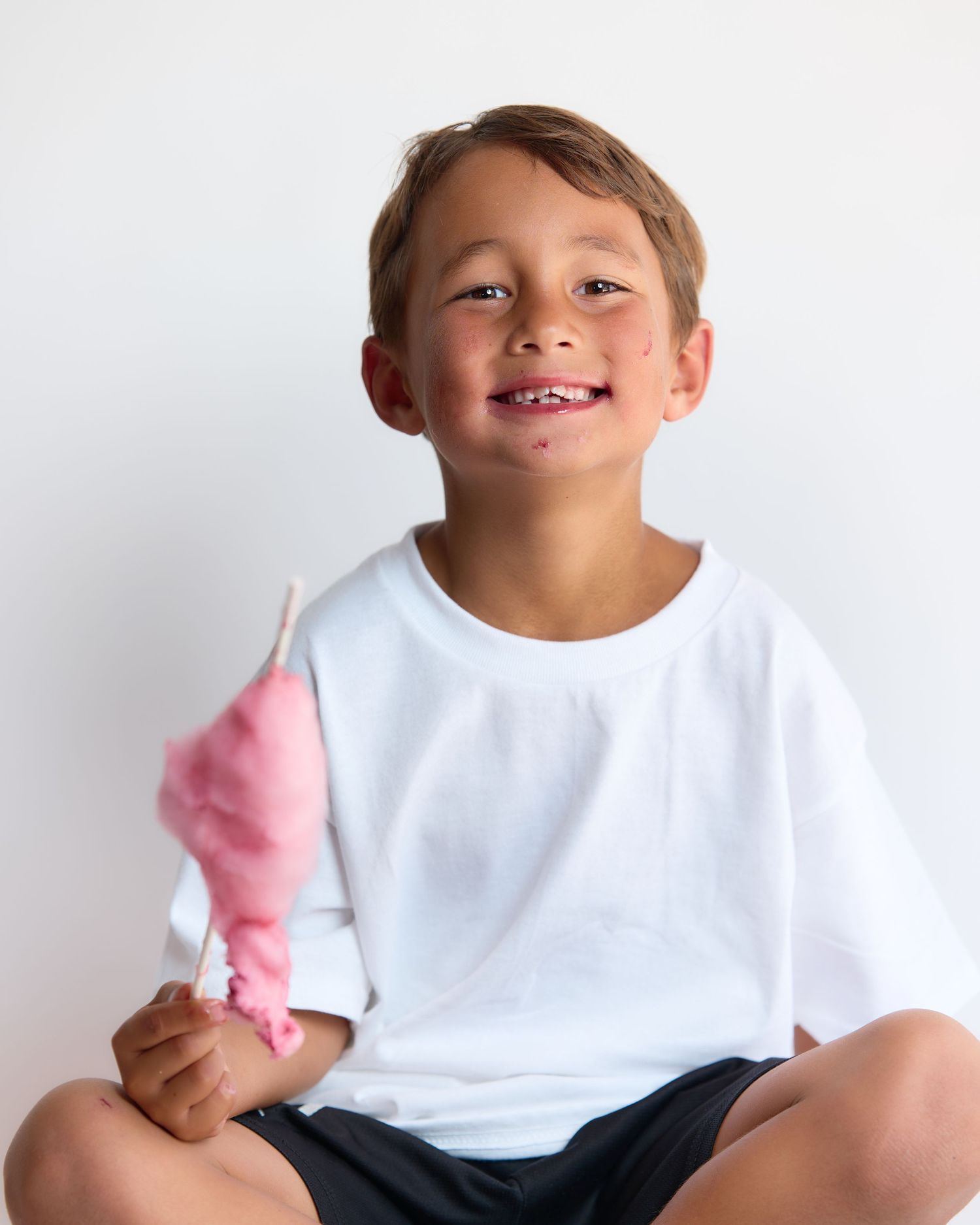 Smiling boy in white t-shirt holding pink cotton candy on a stick, sitting against a white background.