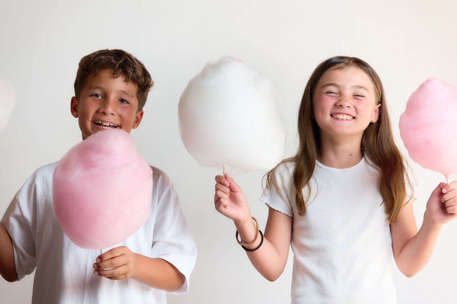 Two happy kids in white shirts holding colorful cotton candy, smiling against a white background.
