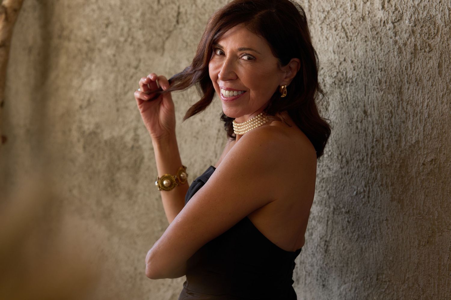 Smiling brunette woman in black top leaning against stone wall, wearing gold jewelry, playfully twirling her hair.