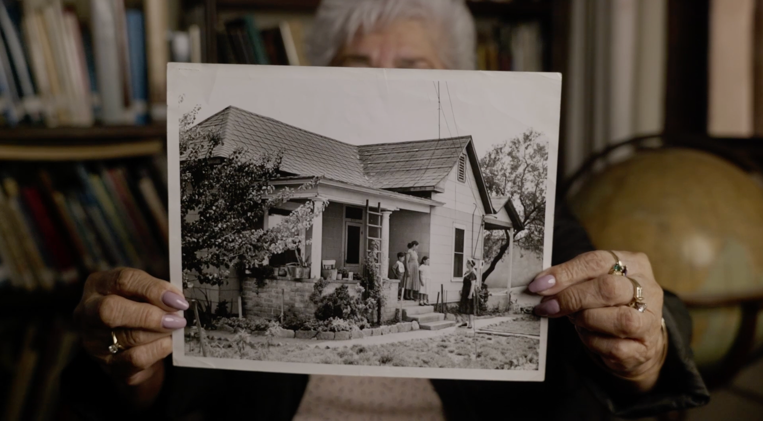 Someone holds an old black and white photograph showing a small house with a front porch and people gathered outside.