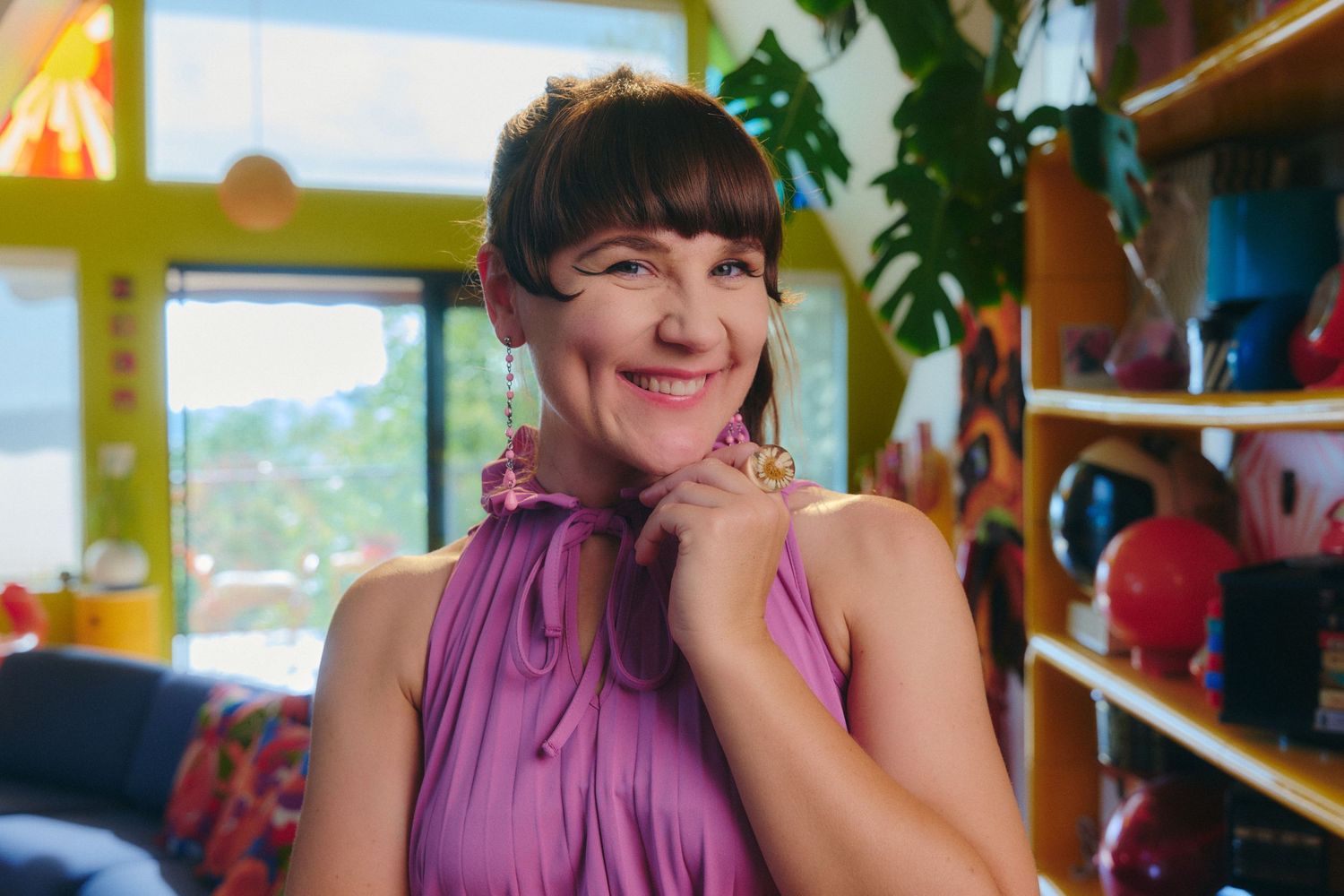 Smiling woman with short dark hair wearing a purple sleeveless top in a colorful, eclectic boutique shop.